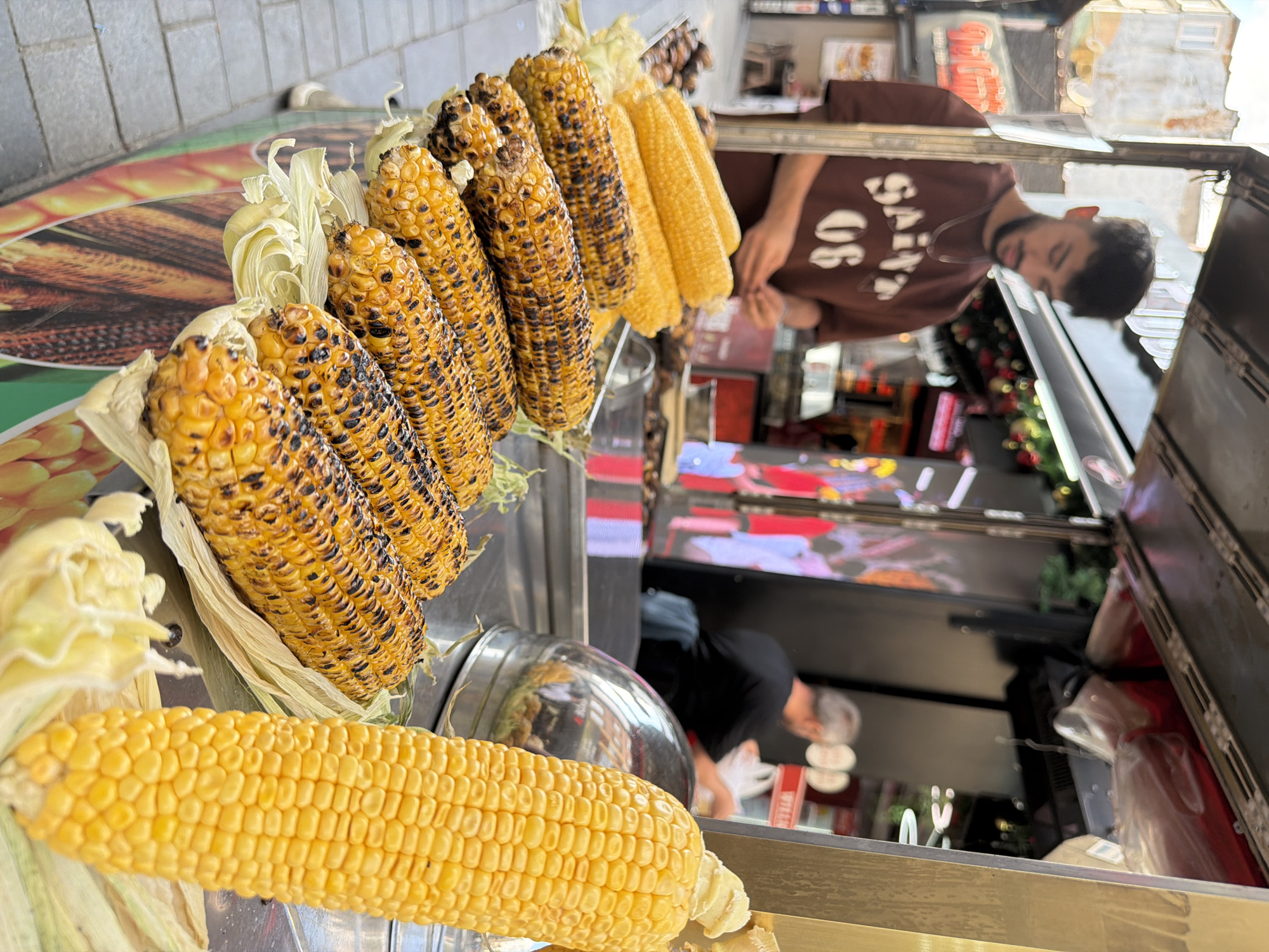 In Kadıköy, Turkey, a street vendor stands behind a cart displaying several ears of grilled corn and unhusked corn. The vendor, a young man with dark hair and a beard, wears a brown t-shirt with a white graphic that reads "SAINT 06". He is looking down at the corn, his hands out of frame, suggesting he is preparing it or interacting with a customer. In the background, another person, an older man with gray hair, is visible inside the stall, facing away from the camera and appearing to be engaged in a transaction. The cart is decorated with images of food. The ground is paved with cobblestones. The scene takes place outdoors, possibly on a street or market. The overall atmosphere suggests a lively urban setting.