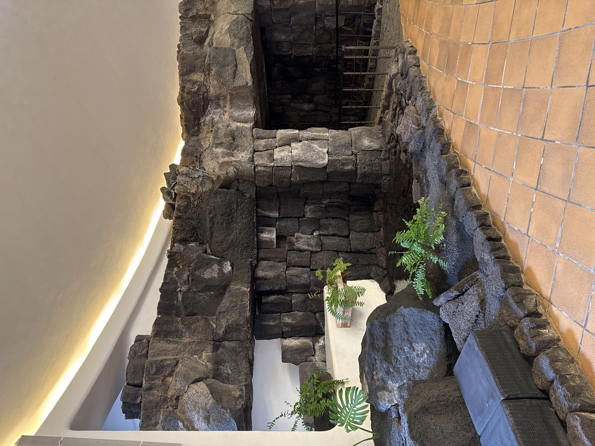 The image depicts an interior space in San Bartolomé, Spain, featuring a structure constructed from dark volcanic rock. The rock forms a multi-tiered arrangement, resembling a natural cave or grotto-like construction, integrated into the building's design. Within the rock structure, there are openings, one of which reveals a partial view of a dark interior with a metal ladder. Plants are strategically placed within the rock formations. One of the plants is a monstera. The building's architecture includes tiled flooring adjacent to the rock structure and a brightly lit white ceiling above. The overall impression is of a space where natural elements are incorporated into the built environment.