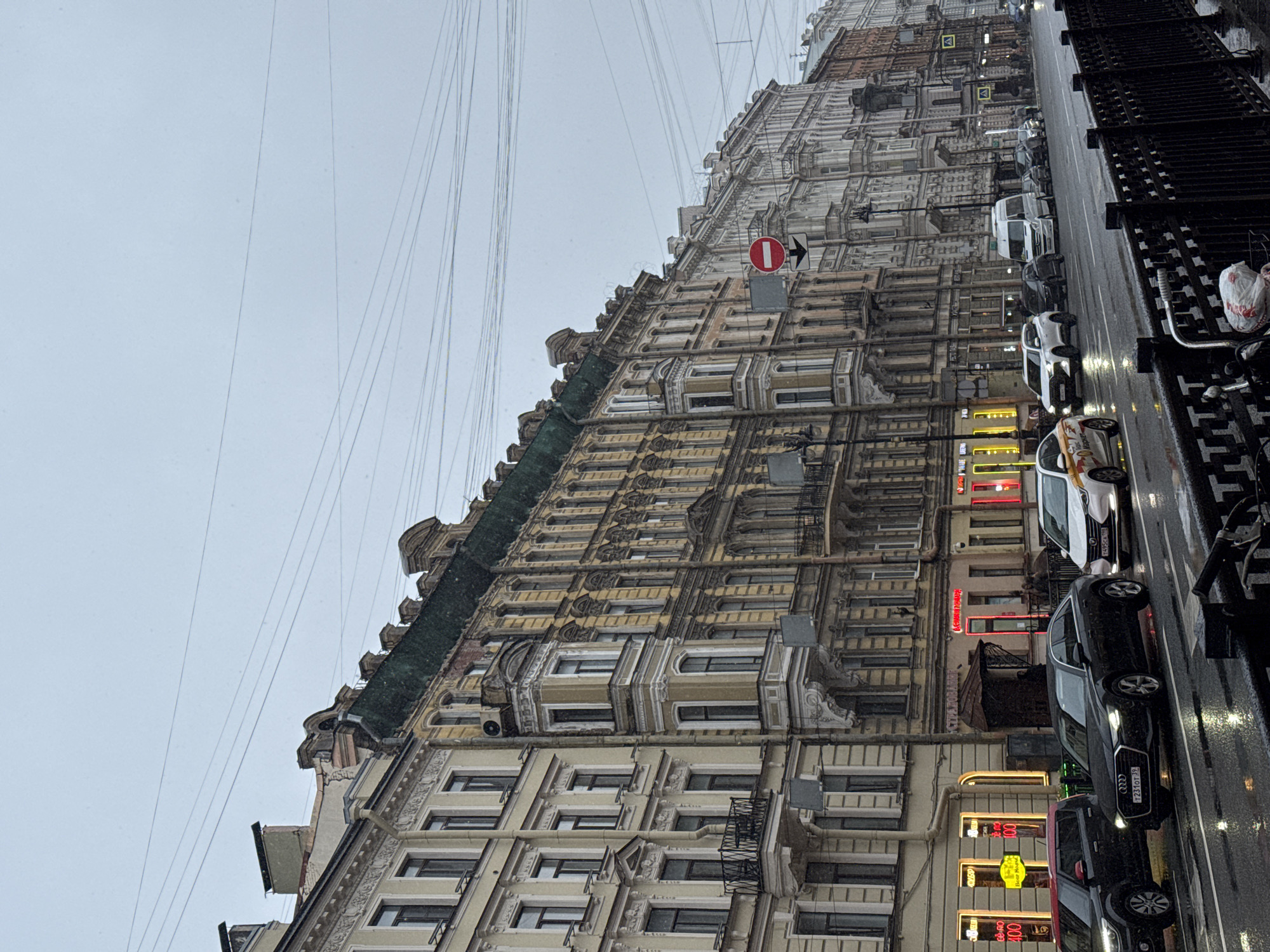 The image, rotated 90 degrees clockwise but described as if correctly oriented, presents a detailed view of a bustling street in Saint Petersburg, Russia, under an overcast, gray sky.
The scene is dominated by a long row of grand, multi-story historical buildings on the left, featuring ornate facades with intricate architectural details, numerous windows, and bay windows. The buildings display varying shades of weathered yellow, beige, and gray. A dense network of overhead wires, likely power or tram lines, crisscrosses against the light sky.
The weather appears cold and damp; the asphalt road is wet and reflective, suggesting recent rain or falling snow, visible as small white specks in the air and on surfaces. Numerous cars are parked bumper-to-bumper along the right curb, including dark-colored sedans and a white van. Russian license plates with regional codes "78" and "178" are clearly visible on several vehicles, confirming the Saint Petersburg location.
Along the ground floor, various storefronts are identifiable by their brightly lit neon signs. Visible Russian text includes "САЛОН КРАСОТЫ" (Beauty Salon) on a red sign, and "ХЛЕБ ПЕКАРНЯ" (Bread Bakery) next to "400" on a yellow sign. A prominent red circular traffic sign with a white horizontal bar and a right-pointing arrow is affixed to one of the upper building facades. On the far right, a dark, decorative metal fence runs parallel to the street. No people are distinctly visible, giving the scene a quiet, active urban atmosphere, characteristic of a cold, perhaps snowy, day.