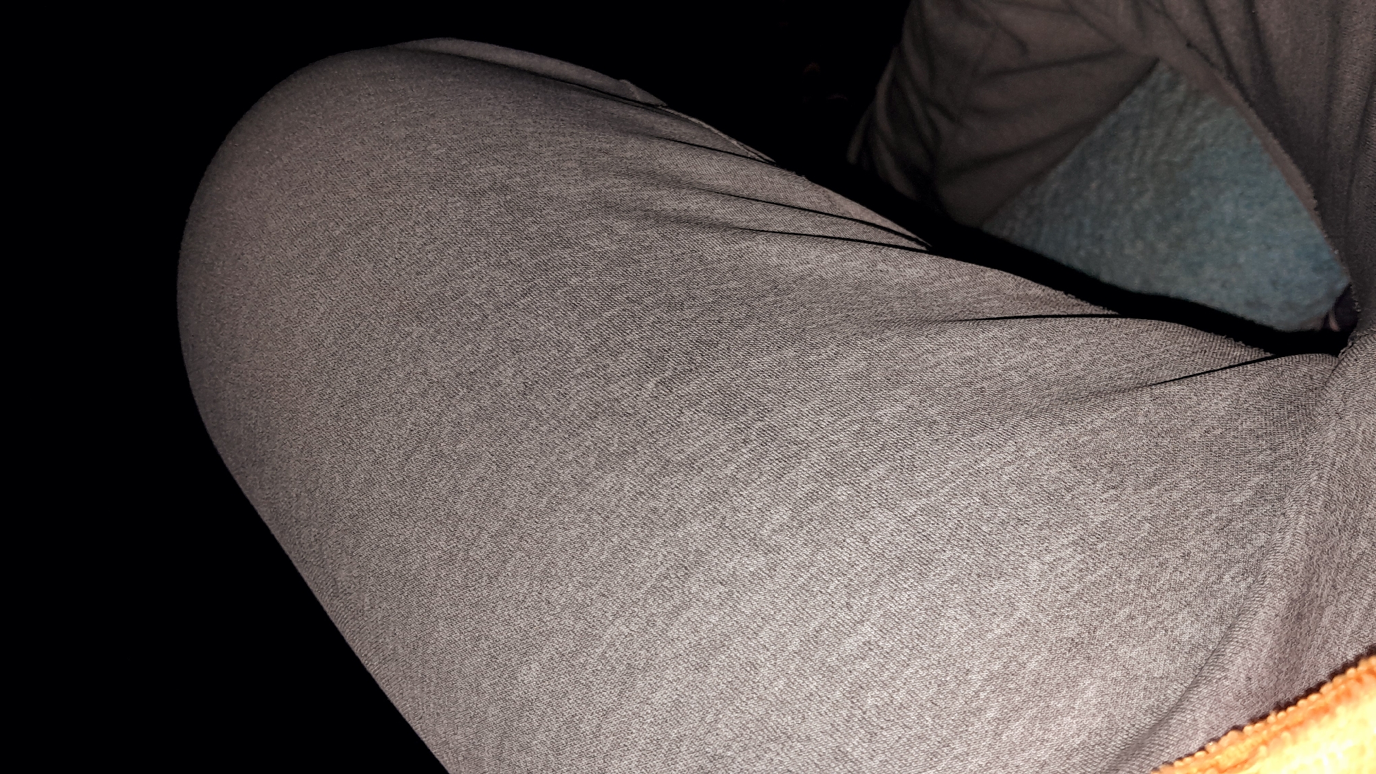 The image displays a close-up view of a person's upper leg, likely a thigh, covered in light grey marled fabric, consistent with sweatpants or casual trousers. The fabric shows a distinct woven or knit texture. The leg appears rounded, suggesting the person is seated or resting.
In the upper right portion of the frame, a darker, possibly olive-green or brown, fabric is visible, which could be part of another garment or a blanket. Beneath this darker fabric and further in the background, a patch of textured, bluish-green fabric, resembling fleece or a soft upholstery, is present, indicating a cushioned surface like a seat or bed. A small fragment of vibrant orange or yellow fabric is just visible at the very bottom right edge.
The scene is characterized by very low lighting conditions, casting most of the surroundings into deep shadows, which suggests an indoor setting or nighttime. Thin, dark lines, possibly seams or elastic bands, run across the grey fabric. No specific activity is taking place other than the person's leg being in this position. There are no discernible location cues to pinpoint the setting to Umuigbekwo, Nigeria, and no visible text is present.