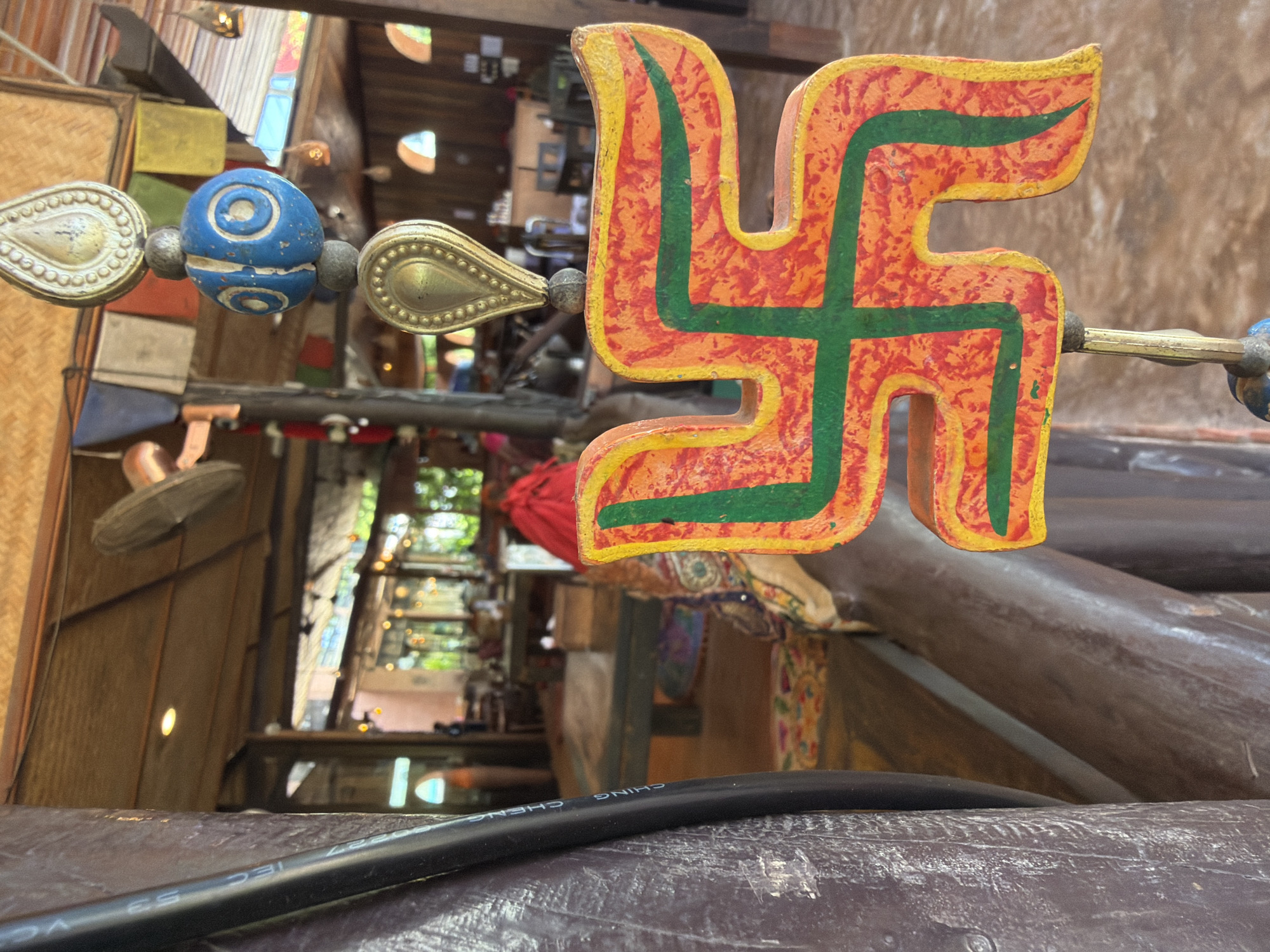 A close-up, eye-level, medium shot features a vibrantly painted, orange and green swastika-shaped charm hanging from a beaded string. The charm is positioned in the foreground, slightly to the right, and is the primary focus. Behind the charm, the scene opens into an indoor-outdoor seating area of what appears to be a restaurant or cafe. Wooden tables and chairs are visible, some with colorful cushions. The background shows a thatched roof structure with exposed wooden beams, and in the distance, blurry figures of people can be seen. The overall ambiance suggests a tropical, relaxed atmosphere, possibly a tourist destination within Koh Phangan, Thailand.