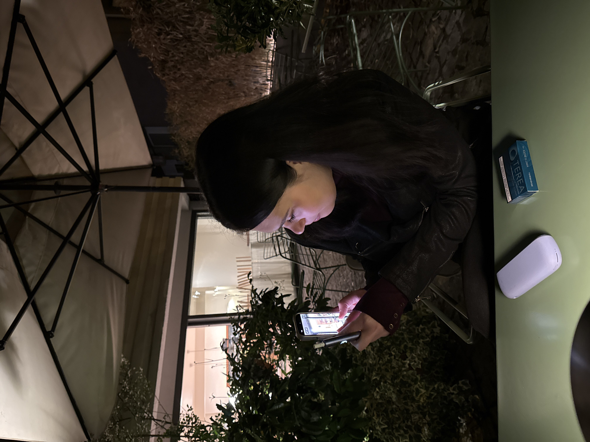 A person with long dark hair, wearing a dark jacket and a dark cap, is sitting at a table outdoors. The person is looking at a smartphone screen and holding a smoking device in their hand. The table is green and has a white pod-like object and a blue package of "TEREA" brand tobacco sticks on it. An umbrella is partially visible above the person. Green foliage and a window are in the background. Based on the "TEREA" product, the scene is likely located in Belgrade, Serbia, where this product is available.