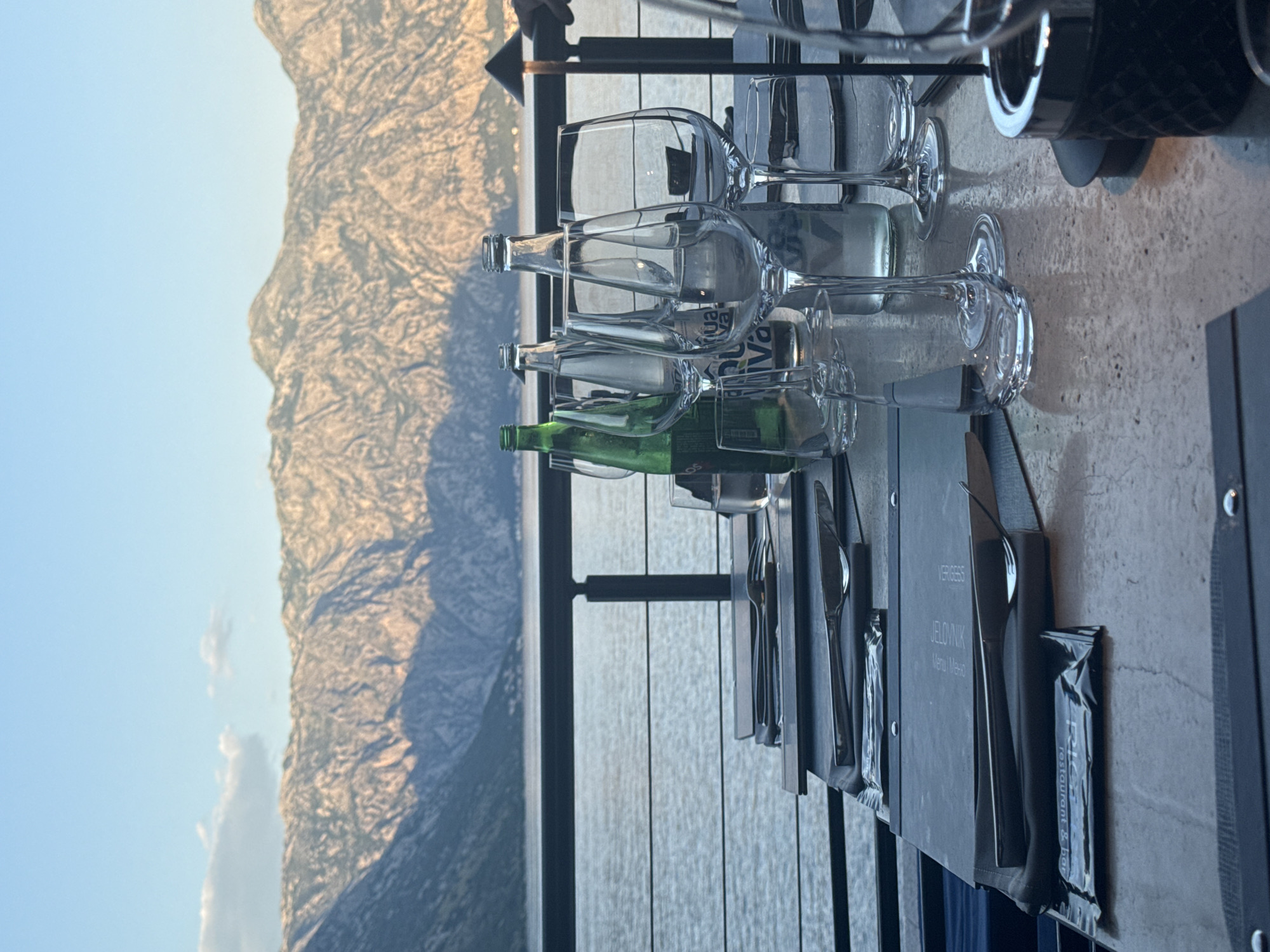 A table is set for dining with a view of mountains and water. There are several clear glass bottles, partially filled with liquid, and stemmed glasses arranged on the table. A green glass bottle is also present. Cutlery and menus are neatly placed on the table next to the glasses. The scene suggests an outdoor dining setting. The location is indicated on one of the menus as Đurići, Montenegro.