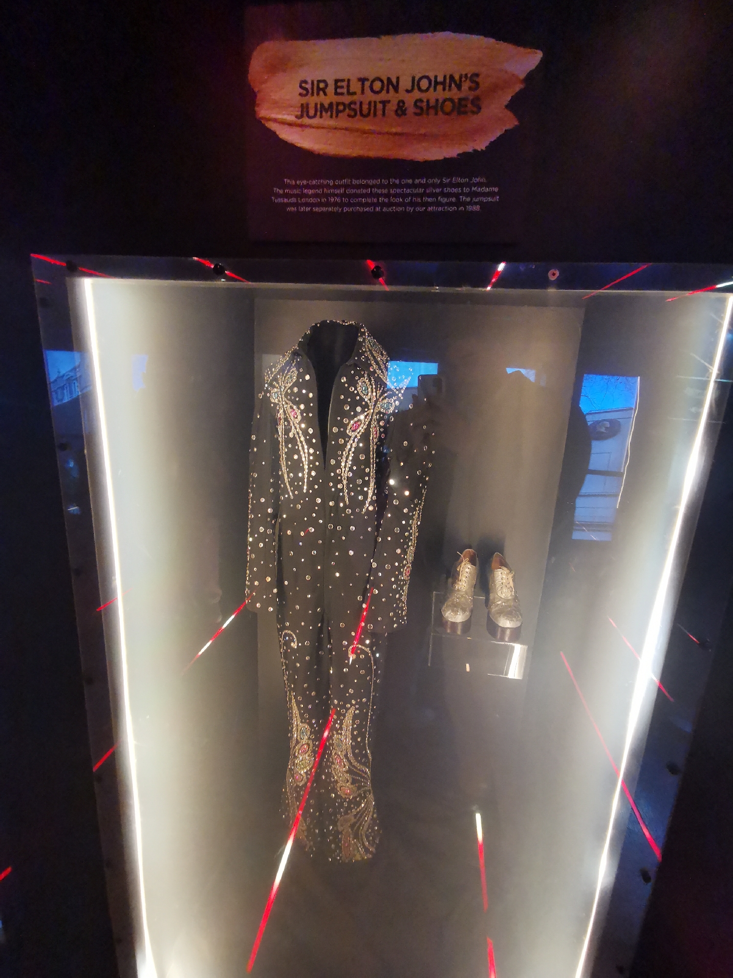 The image shows a display case containing a black jumpsuit and silver shoes. The display is labeled "Sir Elton John's Jumpsuit & Shoes." The text explains that the outfit belonged to Sir Elton John, and he donated the silver shoes to Madame Tussauds London in 1976. The jumpsuit was purchased at auction in 1988 by the attraction, likely implying Madame Tussauds.