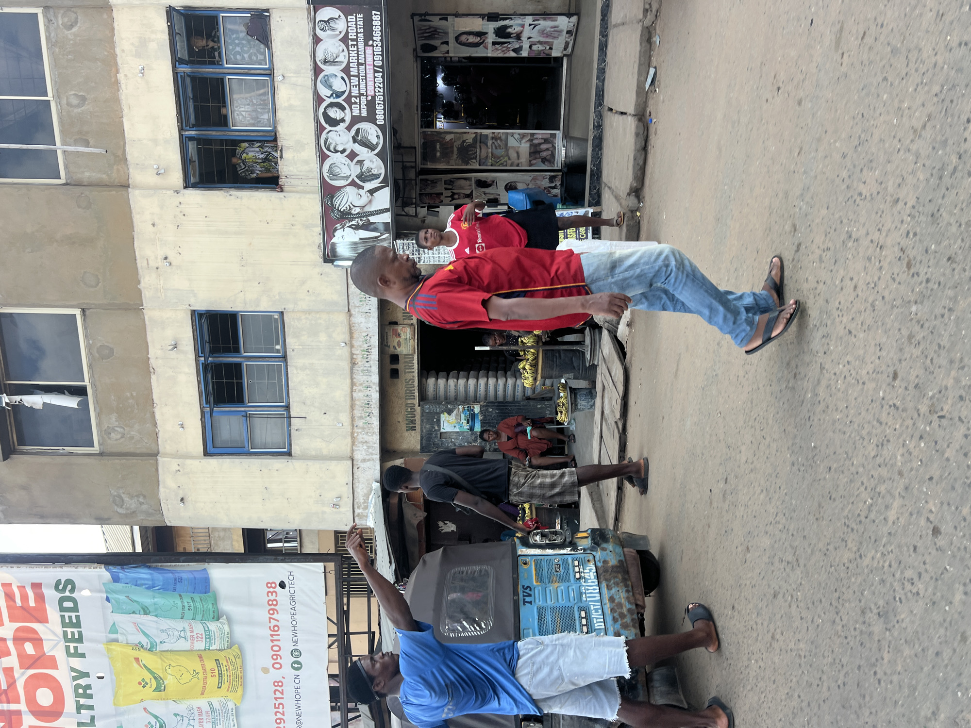 This image captures a street scene in Nkpor, Nigeria. The primary subjects are several men, some of whom appear to be interacting or in transit. In the foreground, a man in a red t-shirt and blue jeans walks towards the right of the frame, his head turned to the left. Behind him, another man in a red t-shirt, possibly a Manchester United fan due to the logo, stands and looks forward. To the left of these men, a large banner advertises "HOPE POULTRY FEEDS" with text detailing different feed types and contact information, including phone numbers like "2925128, 09011679838".
Further into the scene, a blue auto-rickshaw, possibly a form of local transport, is parked. A man in a dark t-shirt and shorts stands beside it, pointing upwards. Another man in a blue t-shirt and white shorts is also near the rickshaw, with his arm raised. In the background, a building with multiple windows and a storefront is visible. A sign above the storefront reads "NWOGO BROS. TRAD." and "NO.2 NEW MARKET ROAD, NKPOR JUNCTION, ANAMBRA STATE", along with contact phone numbers. The storefront appears to be a hair salon, with images of hairstyles displayed. A woman in a red wrapper sits behind the counter.
The overall setting is an urban street with a building on the left and a road in the foreground and middle ground. The lighting suggests it is daytime, likely midday, given the brightness. The activity appears to be normal street life, with people moving about and possibly waiting for or operating transportation. The presence of the banner and storefront indicates commercial activity.