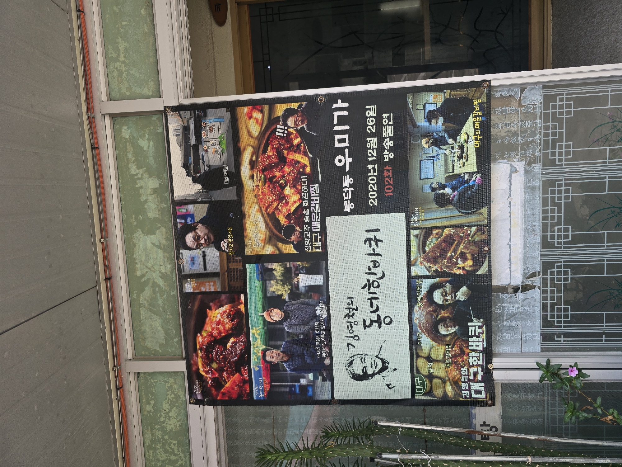The image shows a digital billboard outside a shop in Daegu, South Korea. The billboard displays a grid of images and text promoting a television show or restaurant. The top row features food images and a man with glasses, possibly a host or chef, with text in Korean. The second row shows a couple sitting at a table and another image of food. Below that is a large portrait of a man with Korean text around him, including "Kim Hyung Chul" and "Daegu Han Barbi." The bottom section of the billboard shows another image of people eating at a table with Korean text referring to "Daegu's Spicy Ribs." The billboard is mounted on the exterior wall of a building, with a cactus plant to the left and a window with a traditional lattice design to the right. The overall scene suggests a commercial establishment promoting food or a media program related to local cuisine.