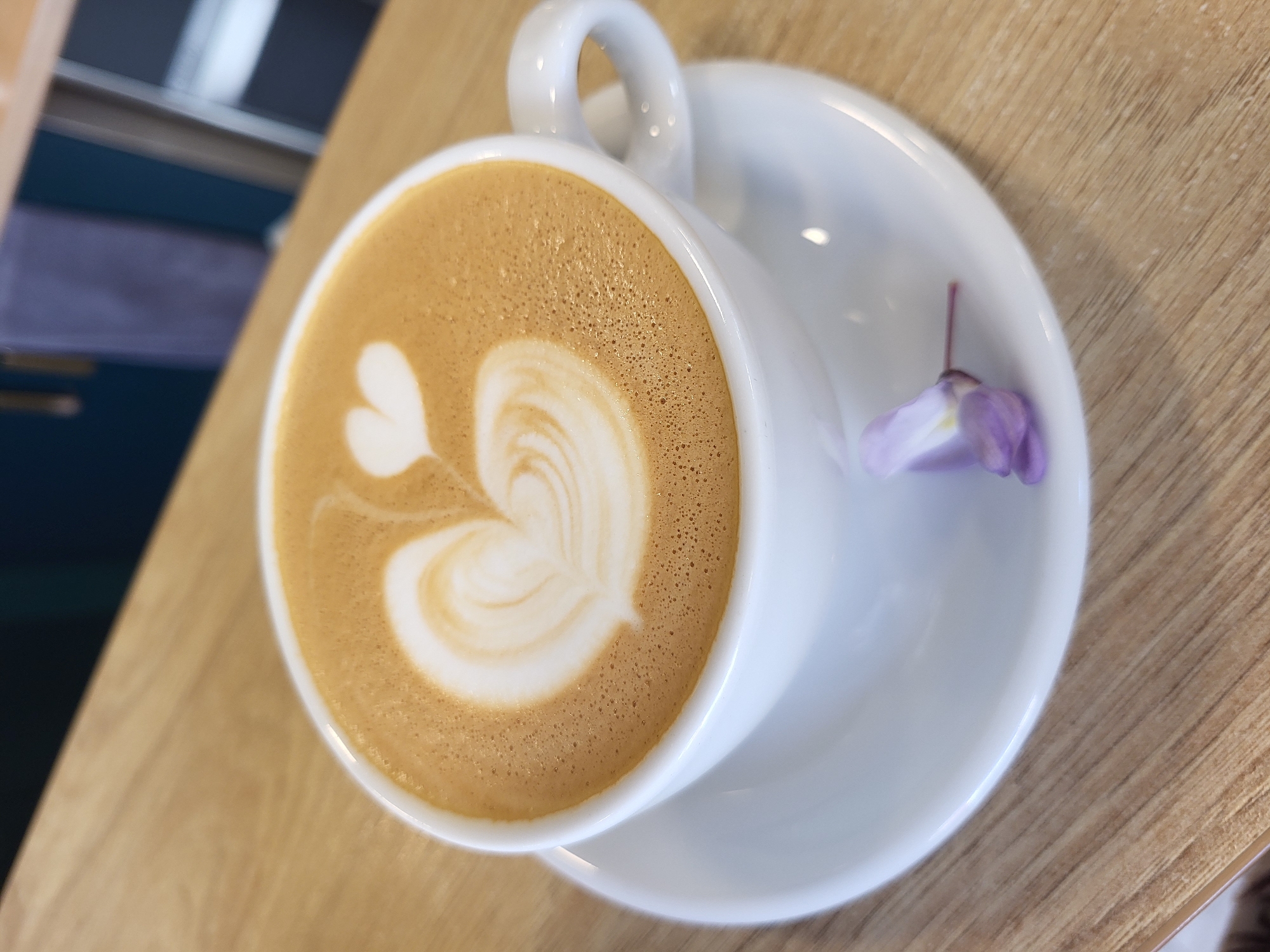 A high-angle close-up shot shows a cup of coffee with latte art placed on a white saucer, which also holds a single wisteria blossom. The coffee features a heart-shaped design in the milk foam. The cup and saucer are set on a wooden table with a light-colored, wood-grain surface. A glimpse of a blue and white surface is visible in the upper left corner of the frame. The location cannot be determined from the image alone.