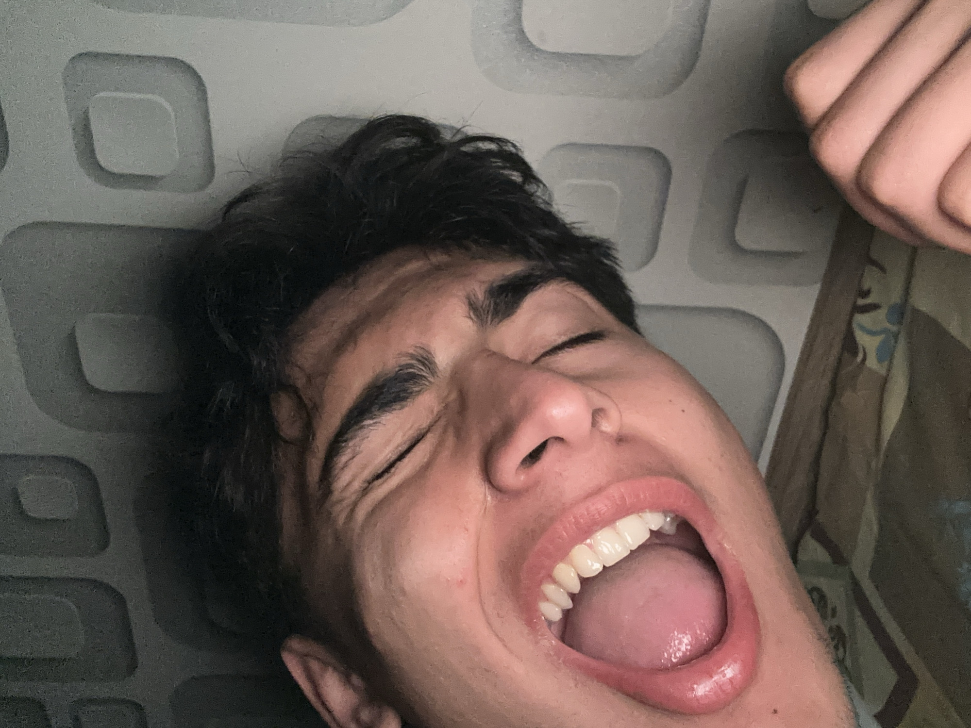 A close-up, low-angle shot shows a young person with dark hair, eyes closed and mouth open in a wide smile or laugh. Their teeth are visible, and the inside of their mouth and tongue are pink. Their skin is light-toned with some subtle pinkness. The background consists of a textured, light gray wall with a repeating pattern of rounded squares. The person is positioned in the lower left portion of the frame, and their head is tilted. A hand with visible knuckles is in the lower right corner, resting on a patterned surface that appears to be bedding. The lighting is dim, creating shadows and highlighting the texture of the wall and the contours of the person's face. The image does not contain any specific visual elements that would allow for the determination of its location in Yalutorovsk, Russia.