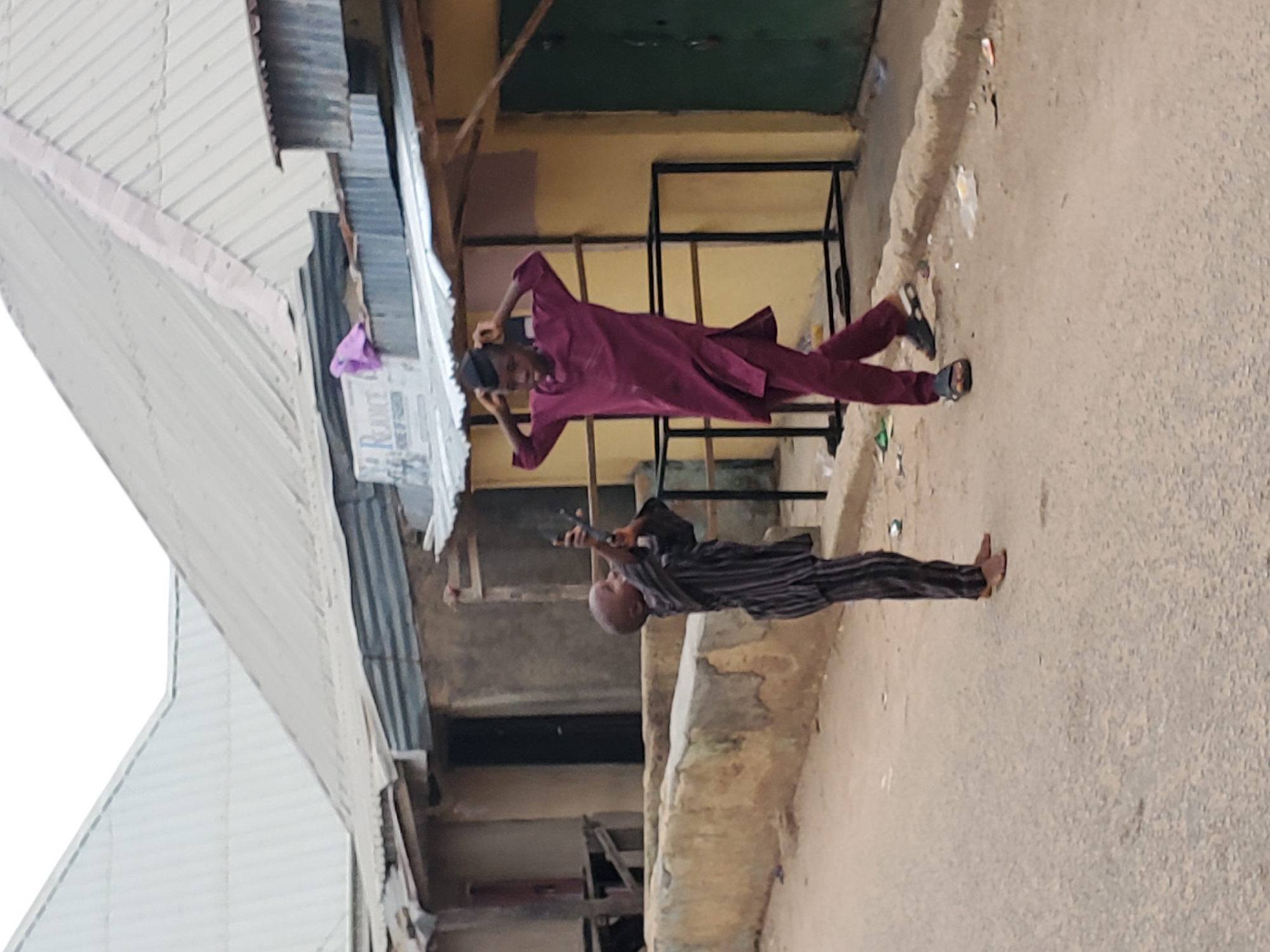 Two men are standing on a street, one is wearing a magenta outfit and the other is wearing a striped outfit. One of the men is holding a phone in the air. The location appears to be Jos, Nigeria, given the architecture and clothing of the subjects. The area appears to be a market or commercial district. The environment includes a mix of permanent and temporary structures.