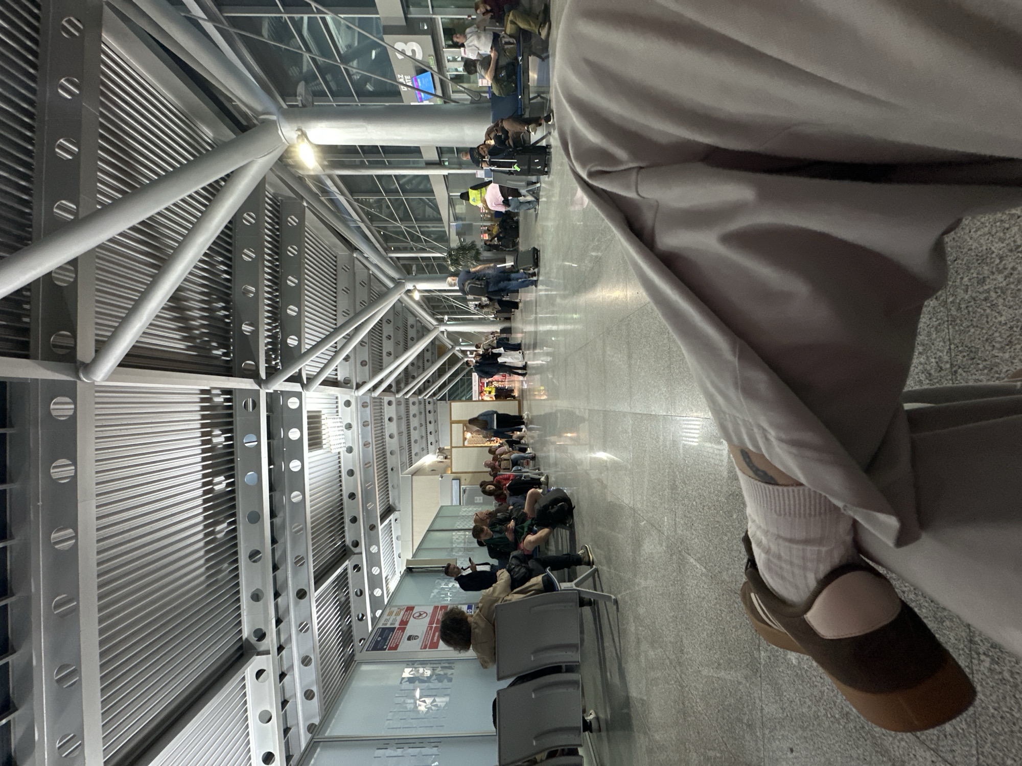 The image shows the interior of what appears to be an airport terminal. People are seated in rows of chairs, likely waiting for flights. The architecture includes a metal ceiling and structural supports, providing a modern and industrial aesthetic. A person is partially visible in the foreground on the right, possibly the photographer. The location is likely Podgorica Airport in Montenegro. The scene is busy, with travelers filling the waiting area.
