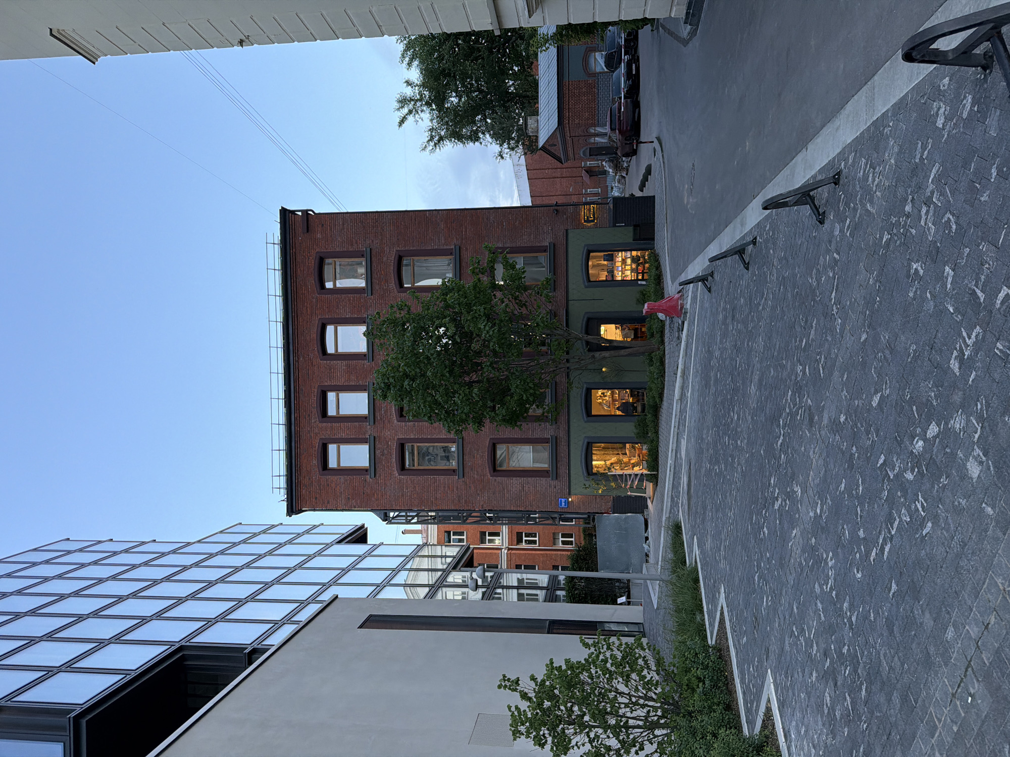 This media file captures an urban street scene in Moscow, Russia, characterized by a blend of old and new architecture. The image is taken from a slightly elevated perspective, looking down into a courtyard or narrow street.
The primary subject is a multi-story, reddish-brown brick building with multiple windows, some of which illuminate interior spaces, suggesting commercial or residential activity. A prominent, dark green lower section of this building also features large display windows, through which interior lighting and merchandise are visible. A small, dark rectangular sign on this green section clearly displays the word "Letters". A deciduous tree stands directly in front of this brick building.
To the left of the brick building, a modern, angular building with a reflective glass facade dominates the foreground, its geometric pattern reflecting the sky. The ground level features a paved area with grey cobblestones or pavers, separated by a white line from an asphalt road or driveway. Several dark metal barriers or street furniture items are installed along the paved area. Patches of green bushes and grass line the edges of the paved surfaces. In the background, beyond the brick building, another structure with brick facade and windows is partially visible, along with what appears to be a parked car.
The sky above is clear and bright blue, indicating good weather. Despite the bright sky, the illuminated interiors of the ground-floor businesses suggest it might be late afternoon or early evening. No people are visible, and no specific event is taking place, portraying a quiet moment in an urban setting.