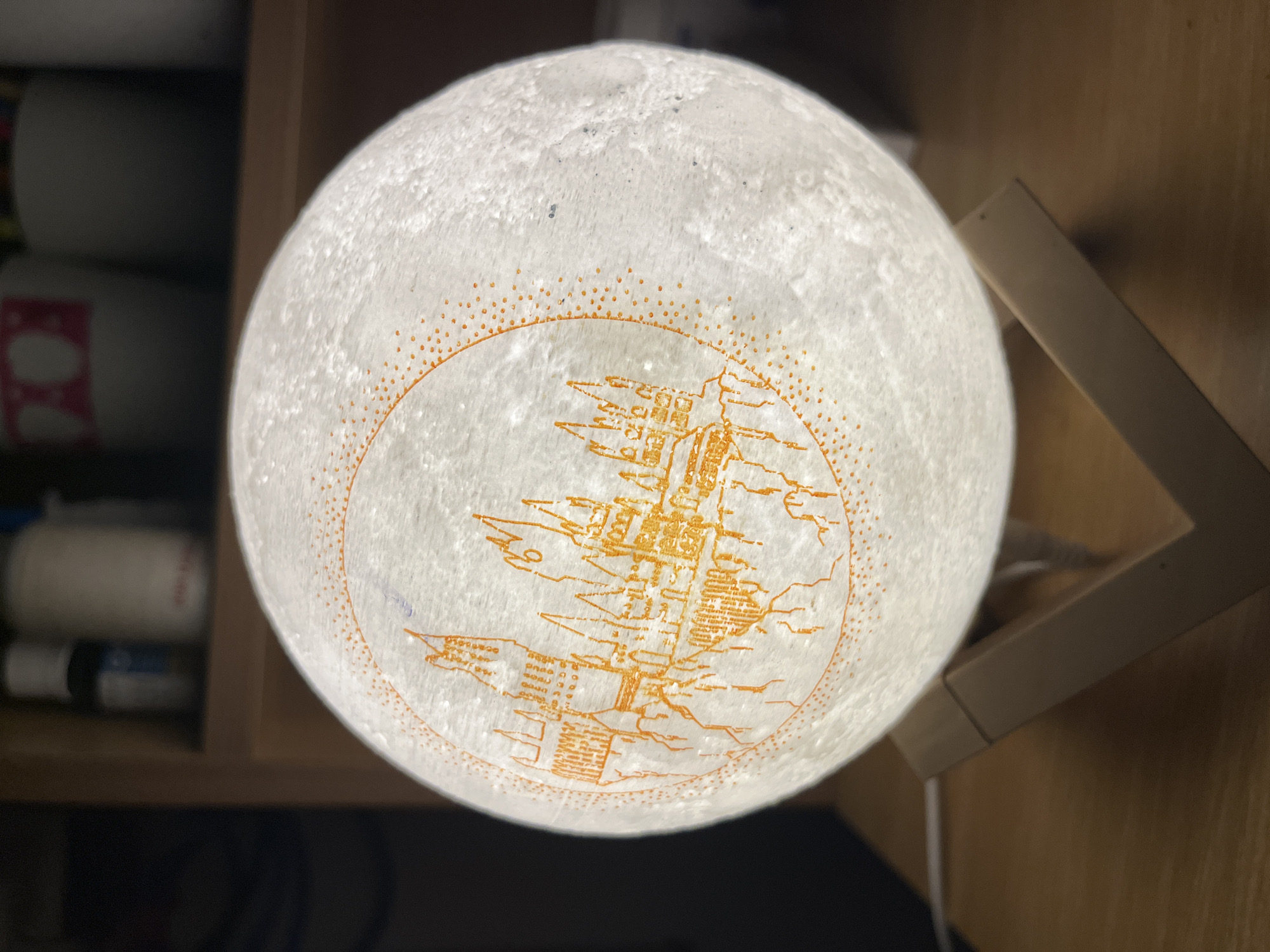 The image shows a spherical lamp resembling the moon, with a textured white surface. A stylized orange drawing is on the surface, depicting buildings or structures. The lamp is placed on a wooden stand with a visible cord. To the left of the lamp are various bottles and containers on a shelf. The location, Bac Nha Trang Ward, Vietnam, cannot be determined from the image.