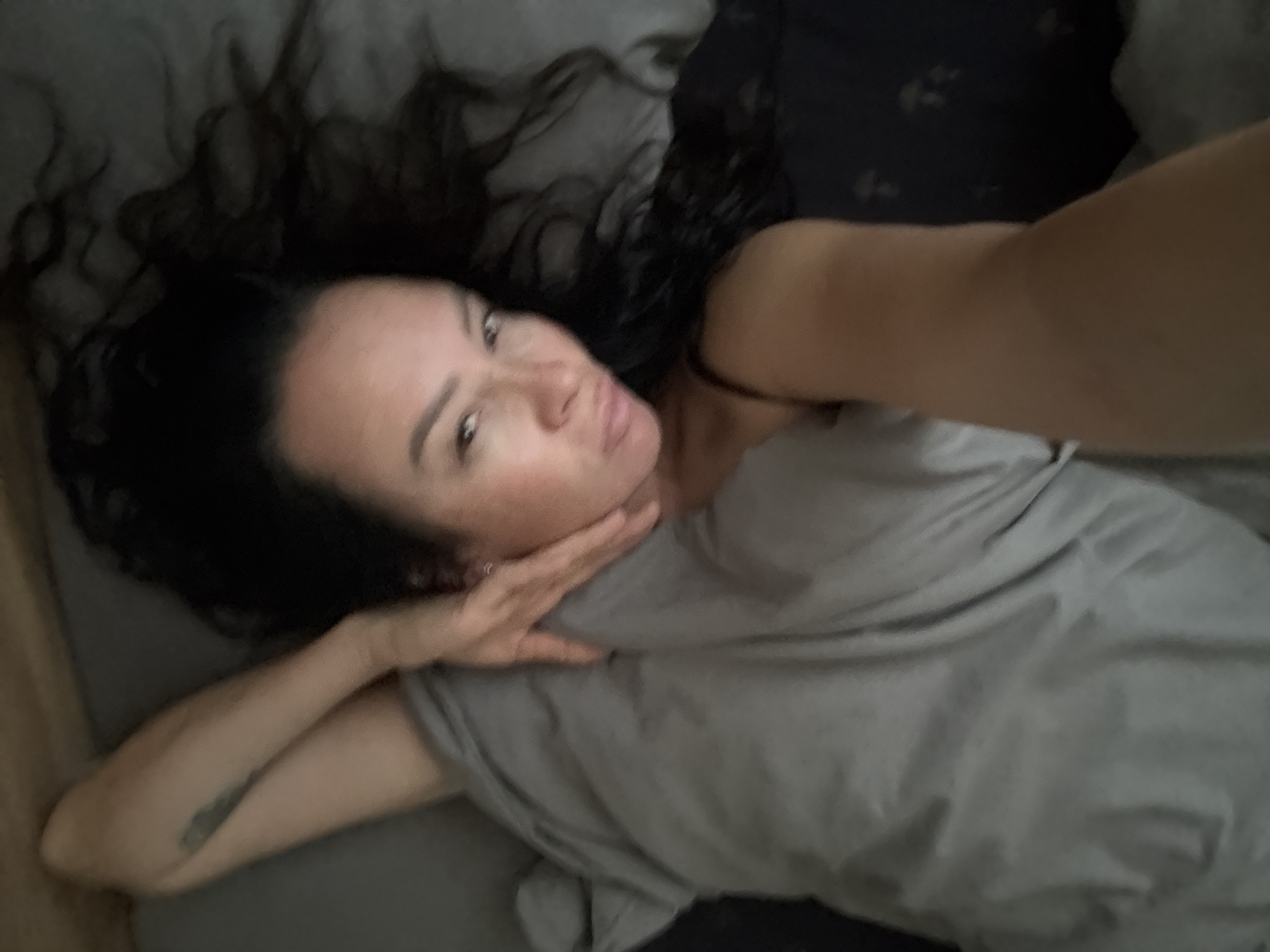 An over-the-shoulder, close-up, low-angle selfie taken from a person lying down on a bed, with their head tilted towards the camera. The person is dark-haired and has freckles on their face. They are wearing a black spaghetti strap top and have a tattoo on their left arm. The bedding appears to be grey. The scene takes place indoors, likely in a bedroom.