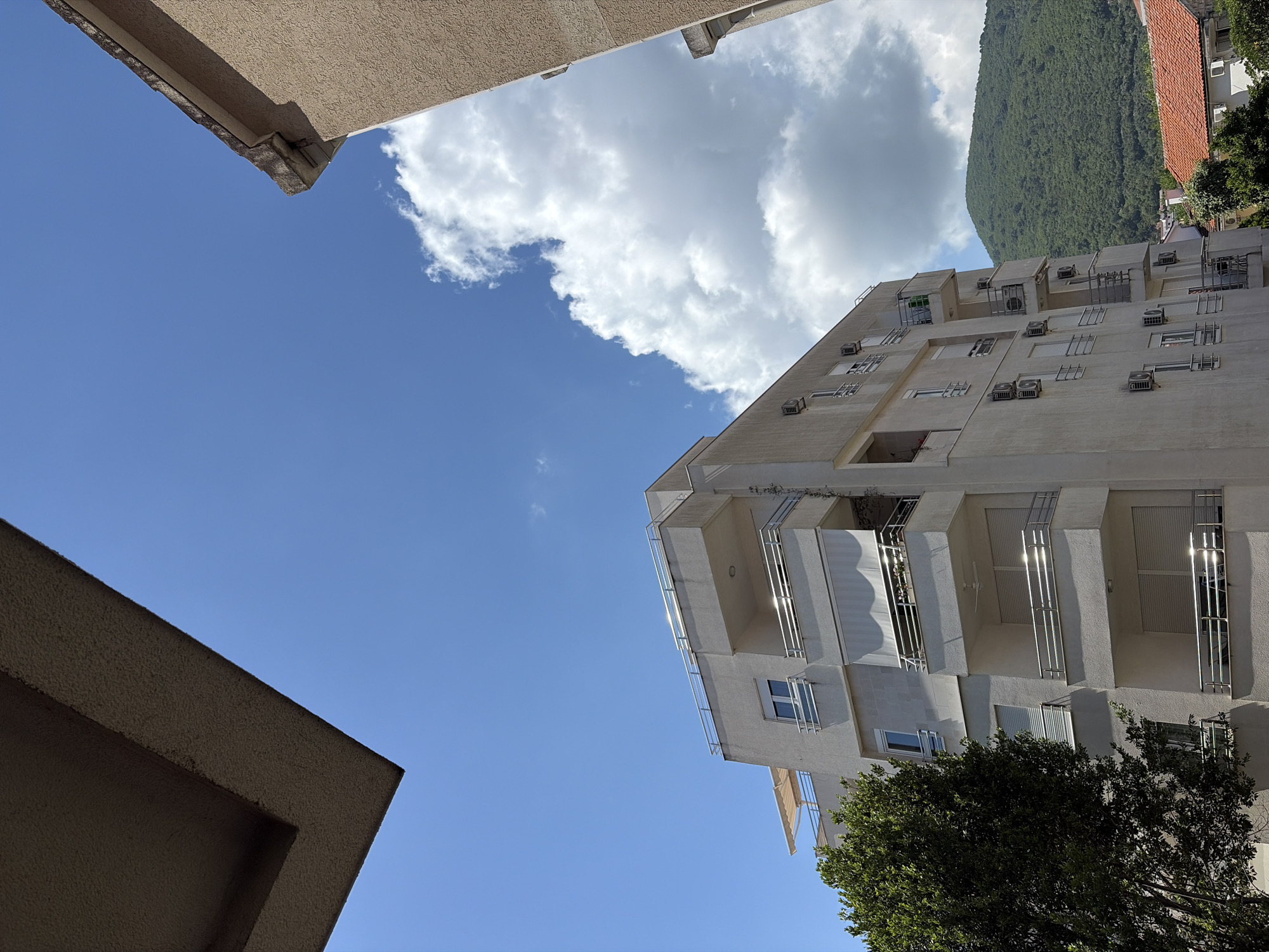The image presents an upward-angled view of an urban landscape under a bright, partly cloudy sky in Budva, Montenegro. A vibrant blue sky dominates the central and left portions of the frame, accented by large, white cumulus clouds.
In the bottom left, the corner of a textured, light brown building partially frames the scene. On the right side, a multi-story, modern apartment building, appearing light beige or white, stands prominently. Its facade is adorned with numerous windows, balconies with metal railings, and external air conditioning units mounted below many windows. Direct sunlight reflects off some of the balcony railings, creating bright highlights.
Behind the apartment building, a lush, verdant green mountain rises steeply, densely covered in trees, suggesting a dramatic natural backdrop to the urban area. A small portion of a building with a red-tiled roof is also visible in the upper right corner, situated between the apartment building and the mountain. A dark green tree with dense foliage partially obscures the lower right section of the apartment building.
The scene is devoid of people and there is no visible text. The bright lighting indicates a sunny daytime setting with clear weather.