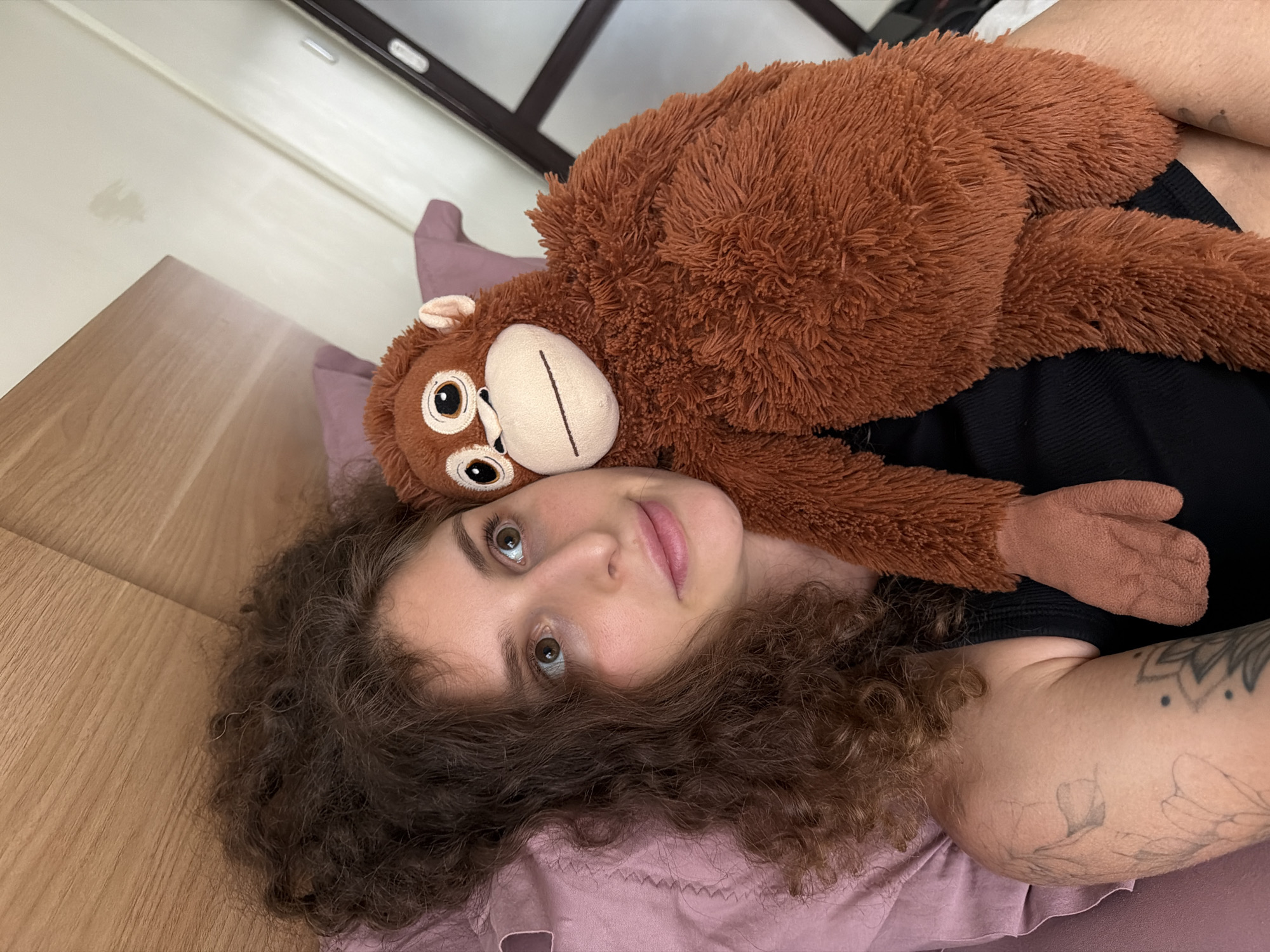 A person with curly brown hair lies in bed with a stuffed monkey. The person is looking upwards with a neutral expression. The stuffed monkey is placed on top of the person's chest and head, with its face visible. The scene is indoors, likely a bedroom, with wooden furniture visible. The lighting suggests it is daytime. The person has tattoos on their arm. The background shows a blurred image of what appears to be a window or mirror, with a dark frame. The overall mood is relaxed and comfortable. The location context is North Kuta, Indonesia.
