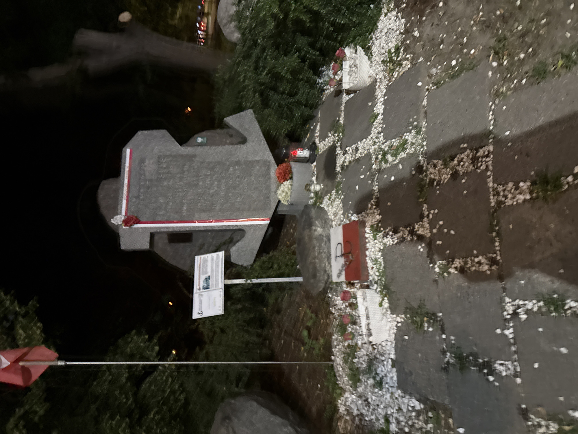 The image shows a memorial at night, located in an outdoor area with trees and foliage in the background. In the foreground, a path made of large stone slabs is covered with white gravel and scattered fallen petals. There are several stone planters containing flowers and candles. One planter to the right holds pink roses in a white woven basket. Another planter to the left holds white and red flowers and a lit candle. A large stone structure, shaped like an anchor, is the central element of the memorial. A Polish flag is planted on a pole to the left of the anchor-shaped monument. A white information board with text and images is mounted on a pole in front of the anchor. Another smaller memorial marker, a red and white plaque with the letter "P" and an anchor symbol, is placed on the ground to the right of the path. The overall scene suggests a commemorative site, possibly related to naval history or a historical event.