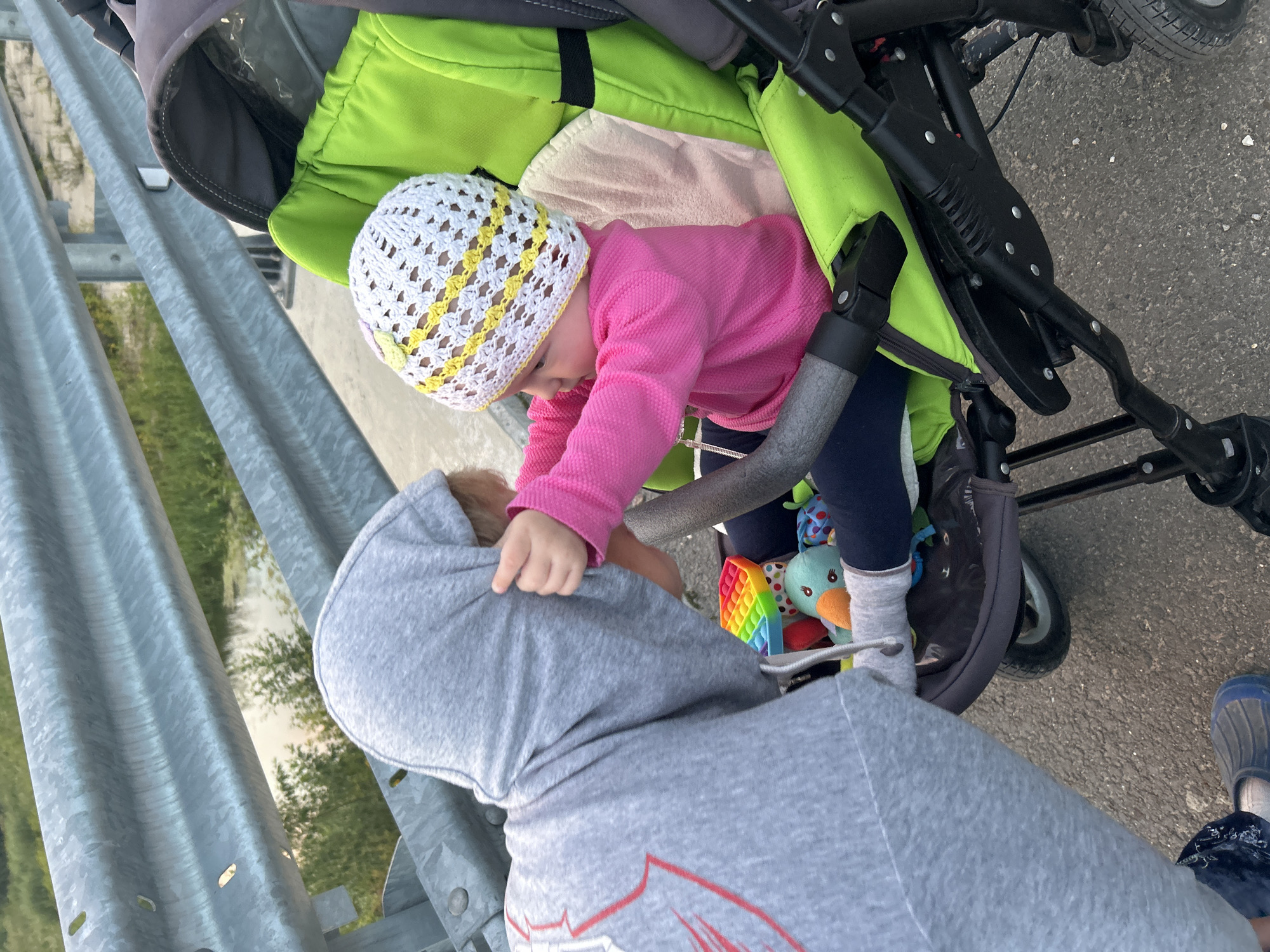 A person wearing a grey hooded sweatshirt is leaning into a stroller, their head obscured by the hood. They are reaching towards a baby seated in the stroller, who is wearing a pink long-sleeved shirt and a white and yellow crocheted hat. The baby is holding onto the sweatshirt. The stroller is green and grey. A colorful pop-it toy and a stuffed bird toy are visible in the stroller. The scene appears to be outdoors, with a metal railing and vegetation visible in the background. The ground is paved. The location is identified as Kalitsino, Russia.