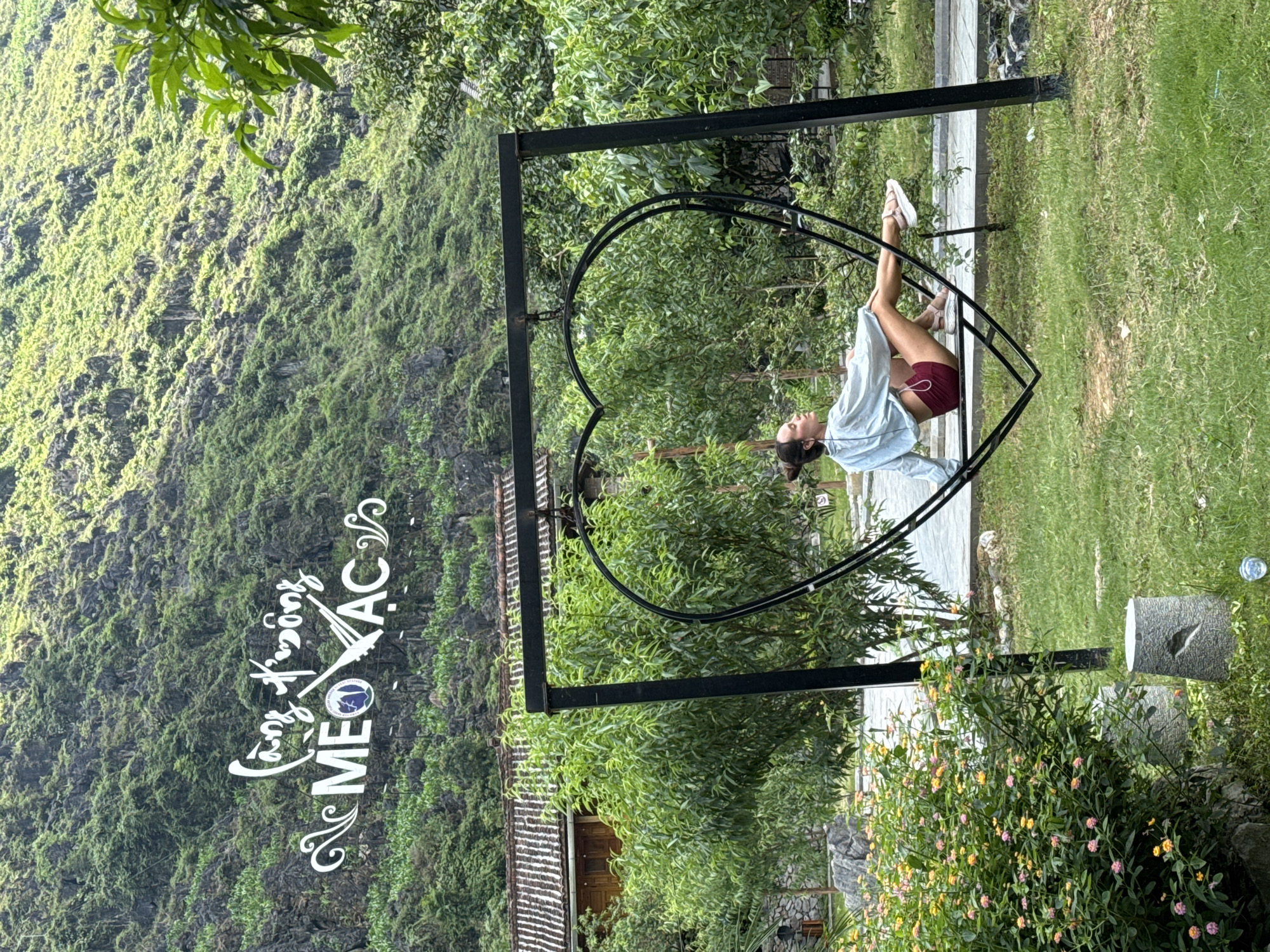 A girl in shorts and a light-colored blouse is resting in a metal heart-shaped swing in Mèo Vạc Commune, Vietnam. Behind the swing are trees and a building. To the left, a steep, mossy rock face rises. A sign with decorative script is on the rock face. The right side of the swing has grassy ground with small, brightly colored flowers. A concrete stump is at the edge of the grassy area.