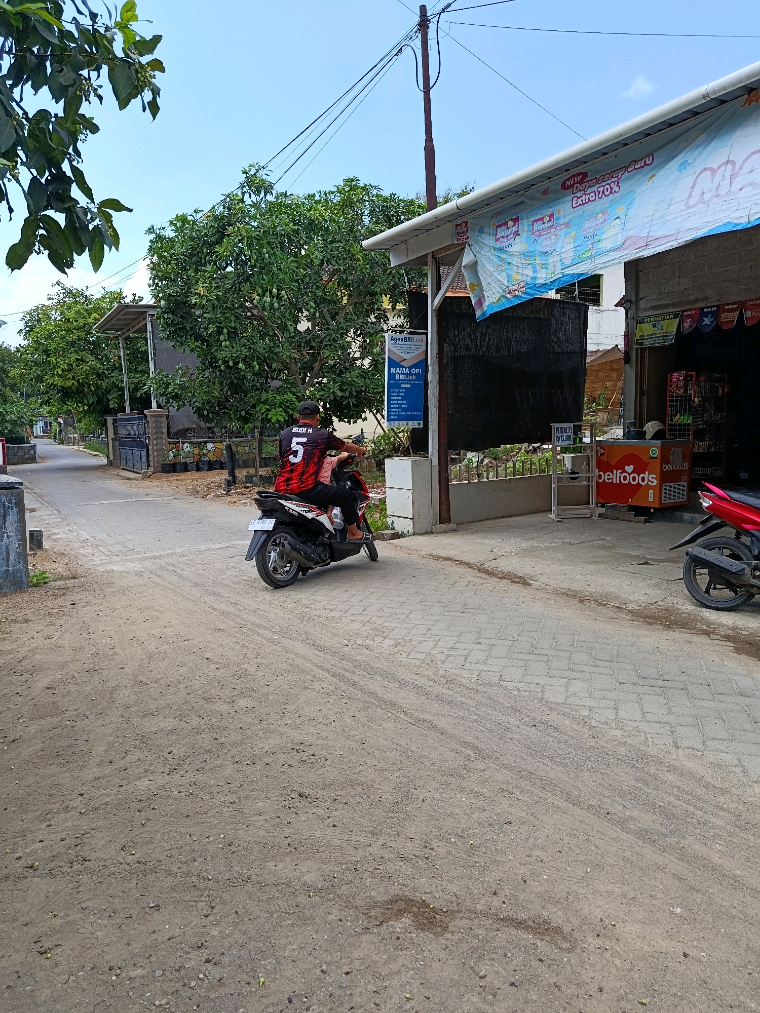 The image depicts a street scene in Menadi, Indonesia, with a focus on a person riding a motorcycle. A person wearing a black and red jersey with the number 5 and the name "BUDI H" is riding a black motorcycle with a child passenger. They are traveling on a dirt road that transitions into a paved walkway. To the right of the road, there is a small shop with a banner displaying promotions and a red motorcycle parked outside. The environment appears to be a semi-urban or rural area, with trees and buildings visible in the background under a clear blue sky. The shop has a refrigerator with the "bellfoods" logo and shelves displaying various items.