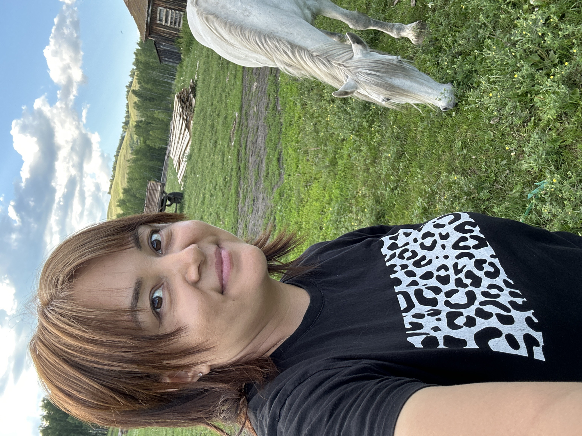 A woman with shoulder-length, reddish-brown hair and bangs takes a selfie in a grassy field. She is wearing a black t-shirt with a white and black leopard print design on the chest. To her right, a white horse grazes in the foreground. In the background, a wooden cart, a pile of lumber, a rustic wooden building, and rolling hills covered in trees are visible under a partly cloudy sky.