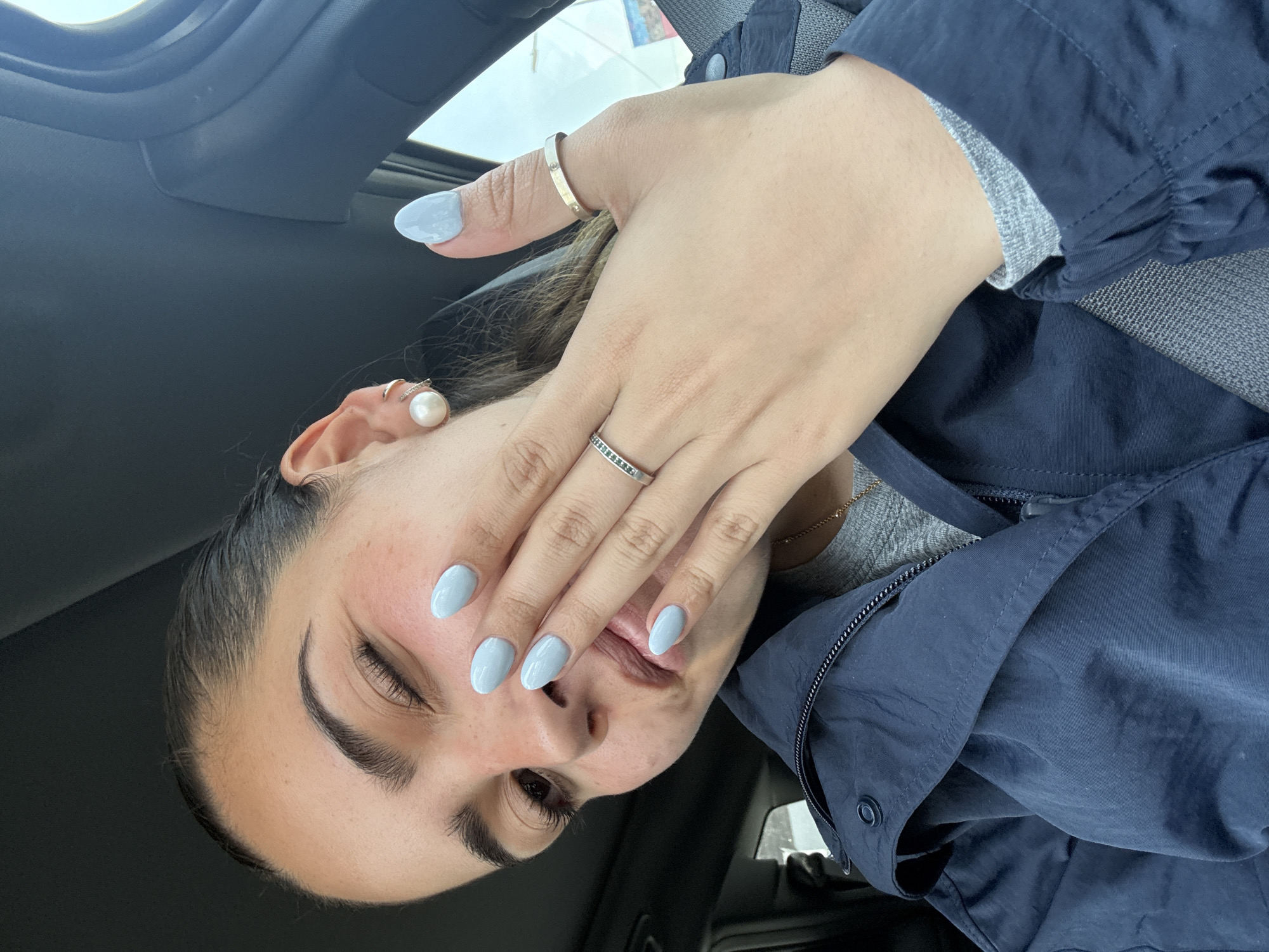An eye-level selfie captured inside a car features a person, likely female, partially obscuring their face with their right hand, creating a playful or coy expression. Their left eye is closed or winking, while the right eye is open and looking towards the camera. The person has dark hair, styled away from the face, and prominent dark eyebrows.
Their right hand, adorned with light blue, oval-shaped nails, covers their mouth and part of their cheek. On the ring finger of this hand, a thin silver-colored ring with small dark stones is visible. Another thicker, plain silver-colored ring is worn on the thumb. A large pearl stud earring and a smaller silver-colored hoop or cuff are visible on the person's left ear. A delicate gold-colored chain necklace can be seen around the neck, peeking out from under their clothing.
The individual is wearing a dark blue jacket, which appears to be made of a slightly crinkled or textured fabric, possibly nylon or similar. Underneath, a grey knit top is visible. The background reveals the interior of a car, with dark upholstery on the seats and ceiling. Bright, diffused light from outside suggests it is daytime, possibly with an overcast sky. The overall impression is casual and intimate, set within the context of Puebla, Mexico. There is no visible text in the image.