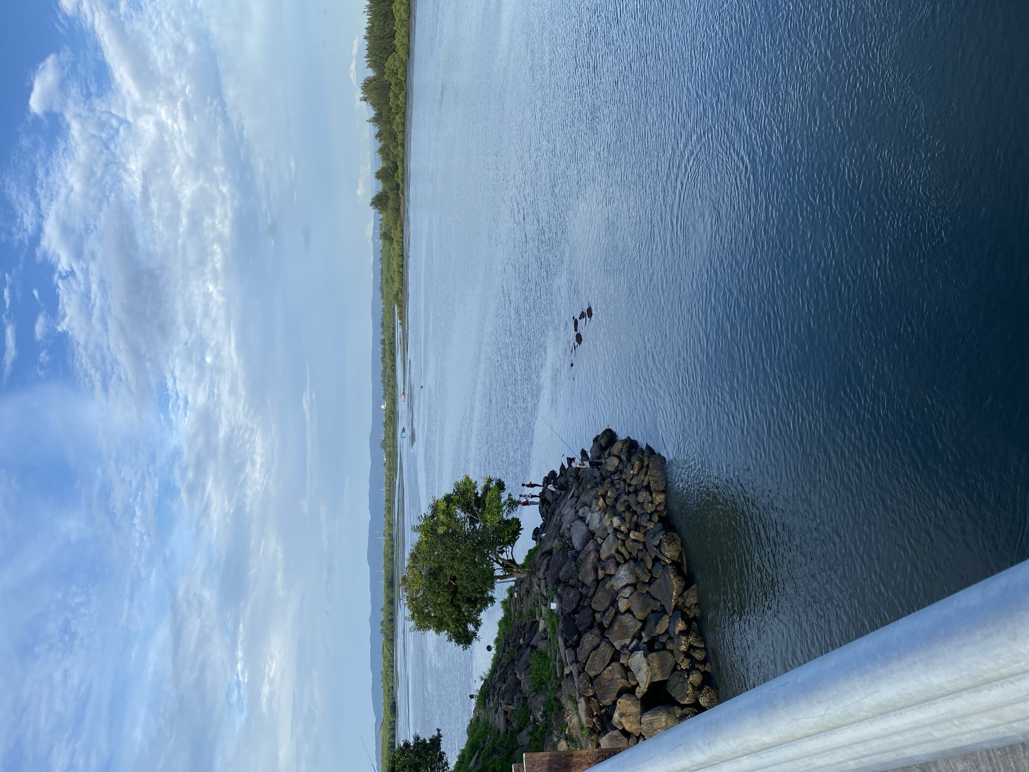 A wide-angle shot captures a serene river scene, likely in Banda Aceh, Indonesia. The foreground features a metallic railing, indicating a bridge or elevated walkway. To the left, a rocky outcrop juts into the water, adorned with a lone, leafy tree. Several individuals are visible on the rocks, some appearing to be fishing. Scattered in the river are a few dark, rectangular objects, possibly buoys or debris. The water is calm and reflects the overcast sky, which is filled with soft, white clouds. The background displays a distant, tree-lined shore and a hazy mountain range.