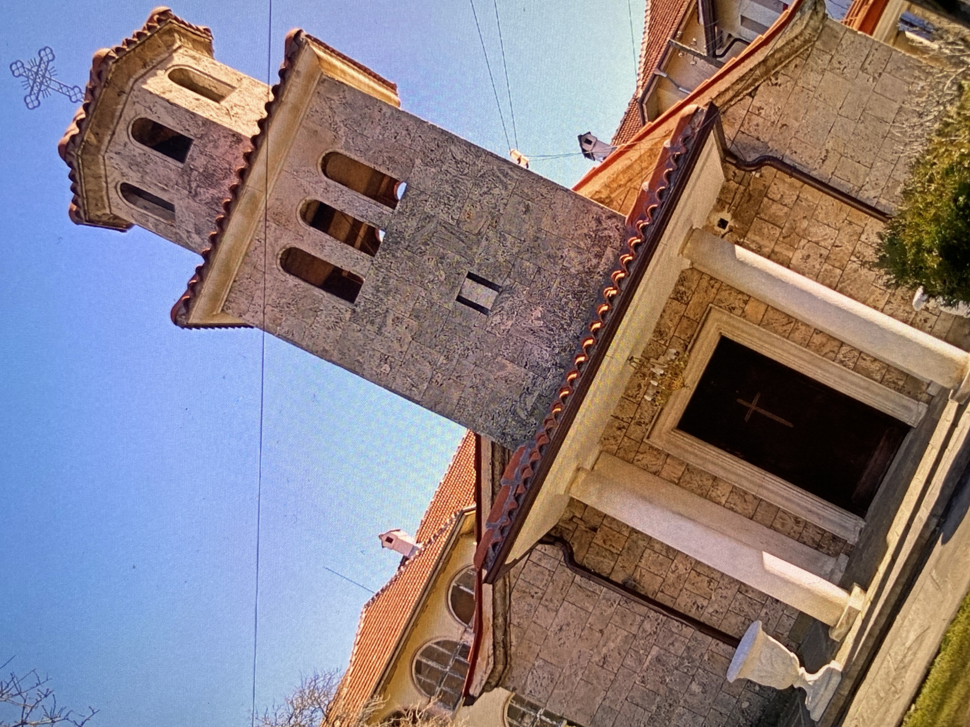 The media file presents an upward-angled view of a stone building, likely a church or monastery, set against a clear blue sky in Bankya, Bulgaria. The image is tilted, showing the structure diagonally.
The primary subject is a sturdy, square bell tower made of light-colored, textured stone blocks. The tower features multiple arched openings towards its top, suggesting a belfry, and is capped with a distinctive, ornate metal cross. The building's roofs are covered with reddish-brown terracotta tiles.
Adjacent to the tower is an entrance area characterized by two prominent white columns supporting a roof overhang. The entrance itself is a dark, recessed doorway, displaying a simple cross design on its surface. Near the entrance, a white, chalice-shaped stone object, possibly a bird bath or a decorative urn, stands on the ground. Further architectural details include round windows with grid patterns on a lower section of the building and a small chimney on one of the tiled roofs.
The scene is captured on a bright, sunny day, indicated by the clear blue sky and visible shadows. There are no clouds, suggesting good weather. Sparse green foliage is visible on the right side of the frame, along with bare tree branches on the left. Overhead utility wires stretch across the sky. No people are present, and no specific activity is taking place. There is no visible text in the image. The overall impression is one of a serene, historically rich architectural structure.