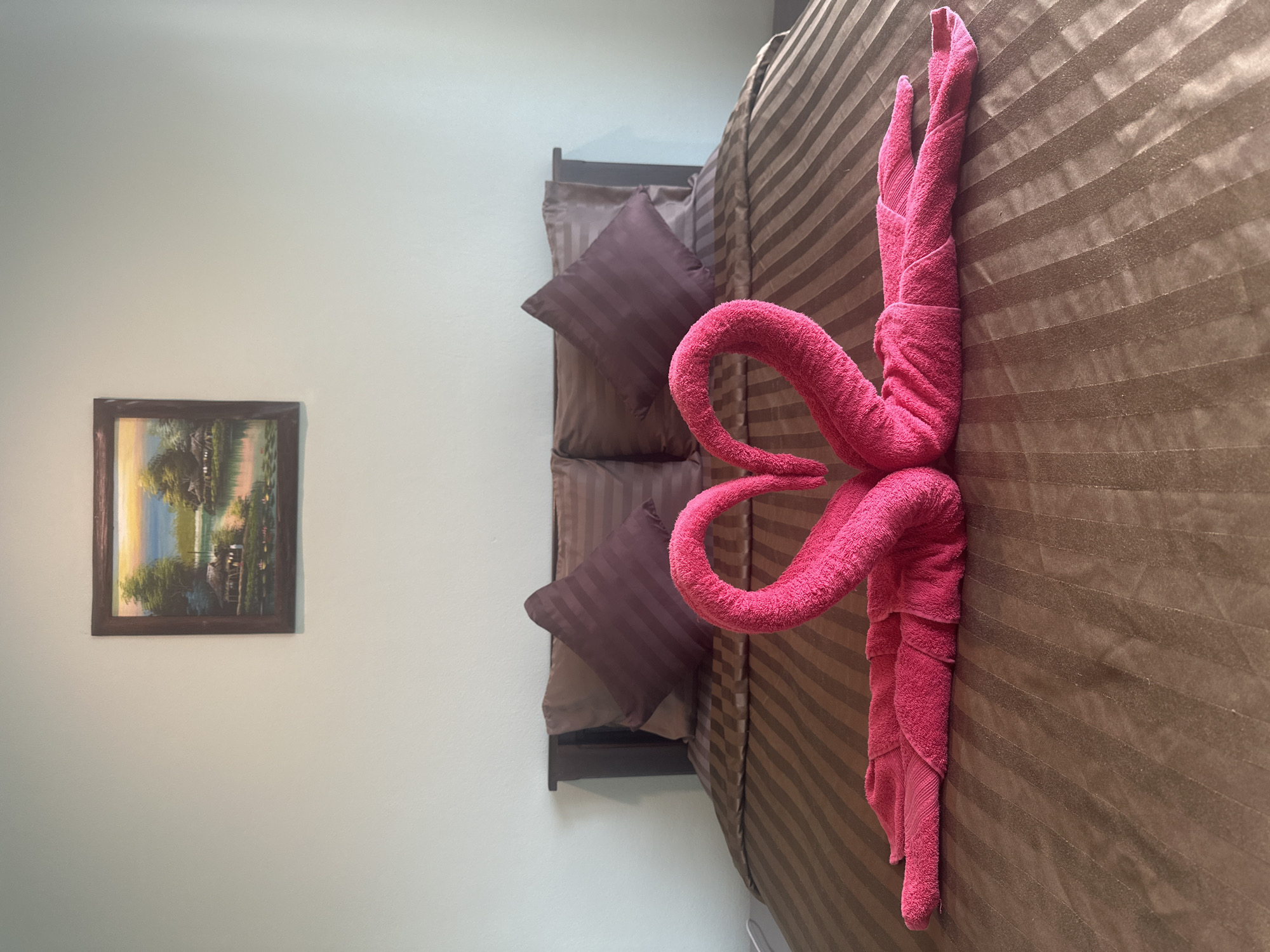 The image displays a bed in a room located in the Ko Samui District of Thailand. The bed is made with brown, striped linens and features two dark brown pillows. In the center of the bed, two pink towels are folded to resemble swans, forming a heart shape with their necks. Above the bed, a framed painting hangs on the wall, depicting a serene landscape with huts by a body of water and lush greenery under a colorful sky. The walls are painted a light, neutral color.
