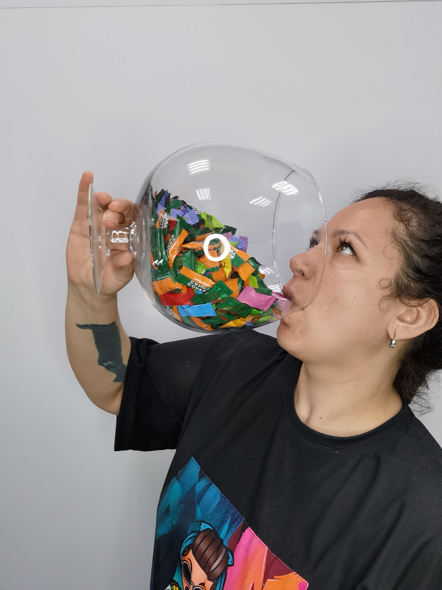 A woman is drinking from a large, clear glass filled with individually wrapped candies. The woman has dark hair pulled back, fair skin, a nose ring, and is wearing a black t-shirt with a colorful graphic design. She has a tattoo on her left forearm. The glass is oversized, resembling a wine glass, and is filled with a variety of candies with colorful wrappers. She is holding the glass up to her mouth and appears to be licking the candies. The background is a plain white wall. The location is Chaikovsky, Russia.