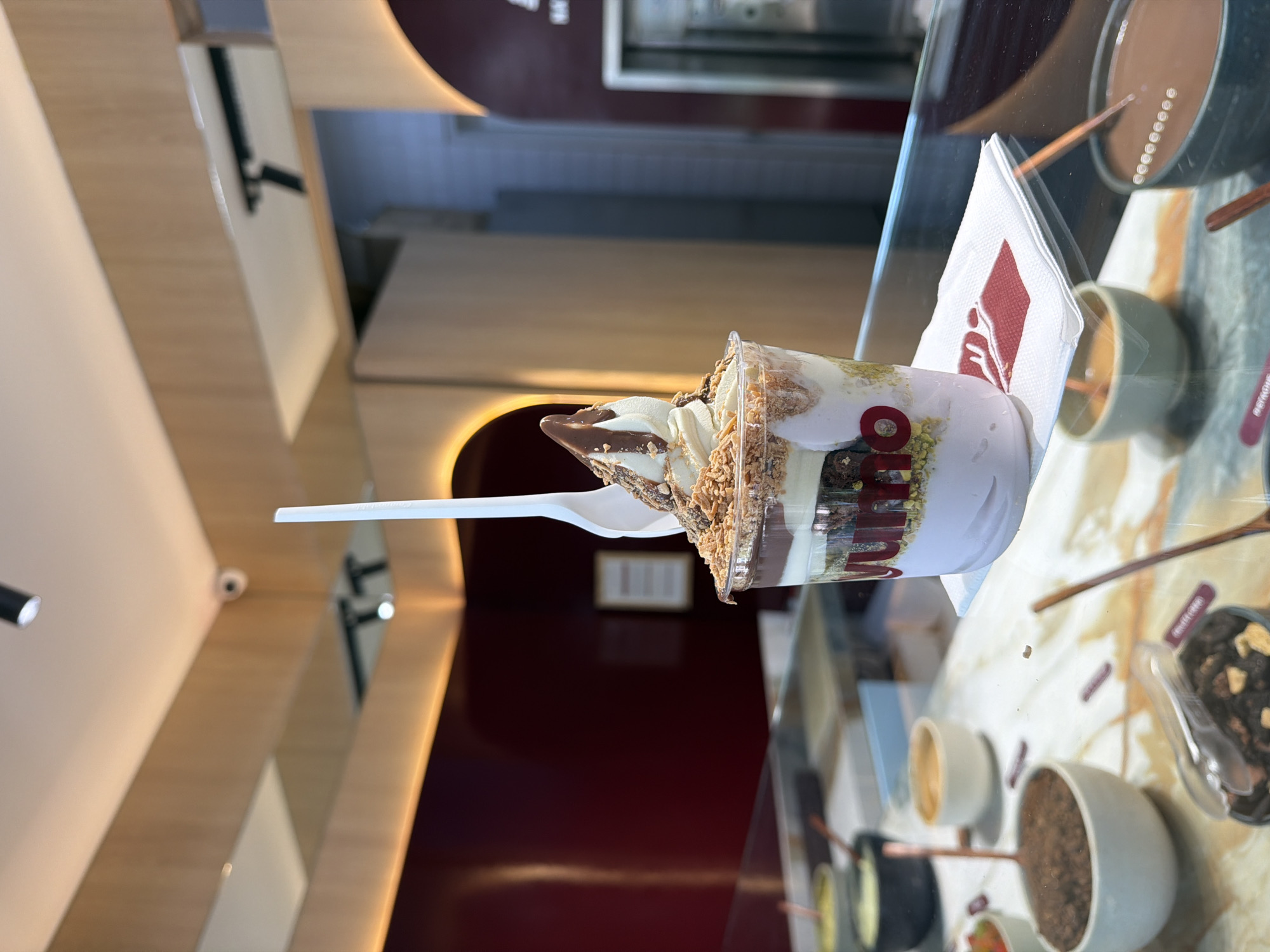 Here is a description of the image:
The image depicts a dessert, likely an ice cream sundae or yogurt parfait, served in a clear plastic cup with layers of ice cream, sauce, and what appears to be crushed nuts and brownies. A white plastic spoon is sticking out of the cup. The dessert is placed on a white napkin with a red logo on a countertop, likely within a dessert shop display case. The countertop is cluttered with small bowls containing various toppings. The background suggests a modern interior with burgundy walls, wooden details, and stainless steel accents. The location is in Mexico City, Mexico, as evidenced by the "Milo" logo on the cup.
