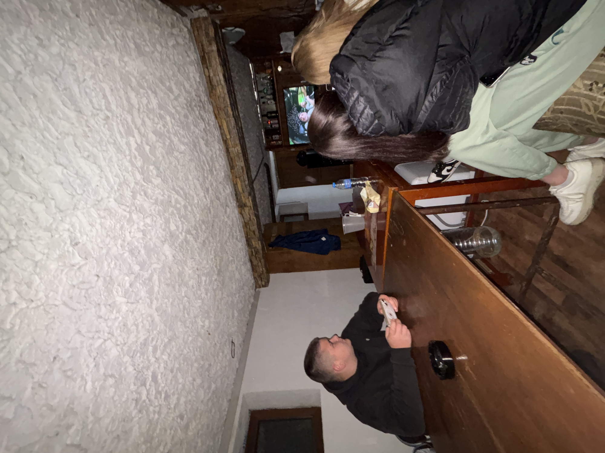 This image captures an indoor social gathering in Lehchevo, Bulgaria, likely during the evening or night. The scene is set in a room with distinctive textured white walls on the left and various wooden elements, including a large, dark wooden table or bar counter dominating the right side of the frame.
Three individuals are present. In the foreground, a man with short hair, wearing a black hoodie, is seen in profile, leaning on the wooden table. He is focused on a white smartphone held in his hands. A black ashtray is also visible on the table near him. Further back, two women are partially visible behind the table, appearing to watch a television screen mounted on a wall in the background. One woman has long dark brown hair and is wearing a black puffer jacket, while the other, with long blonde hair, is dressed in light green sweatpants and white sneakers. Another pair of black and white sneakers is visible on the floor beneath the table.
The room features a television displaying what appears to be people or a video. Behind the TV, shelves hold several bottles, possibly indicating a bar or beverage area. A plastic water bottle is also on the table. On the floor next to the table, a shisha (hookah) pipe with a glass base is visible, along with a dark blue piece of clothing. The overall atmosphere suggests a casual and relaxed social event. No discernible text is visible within the image.