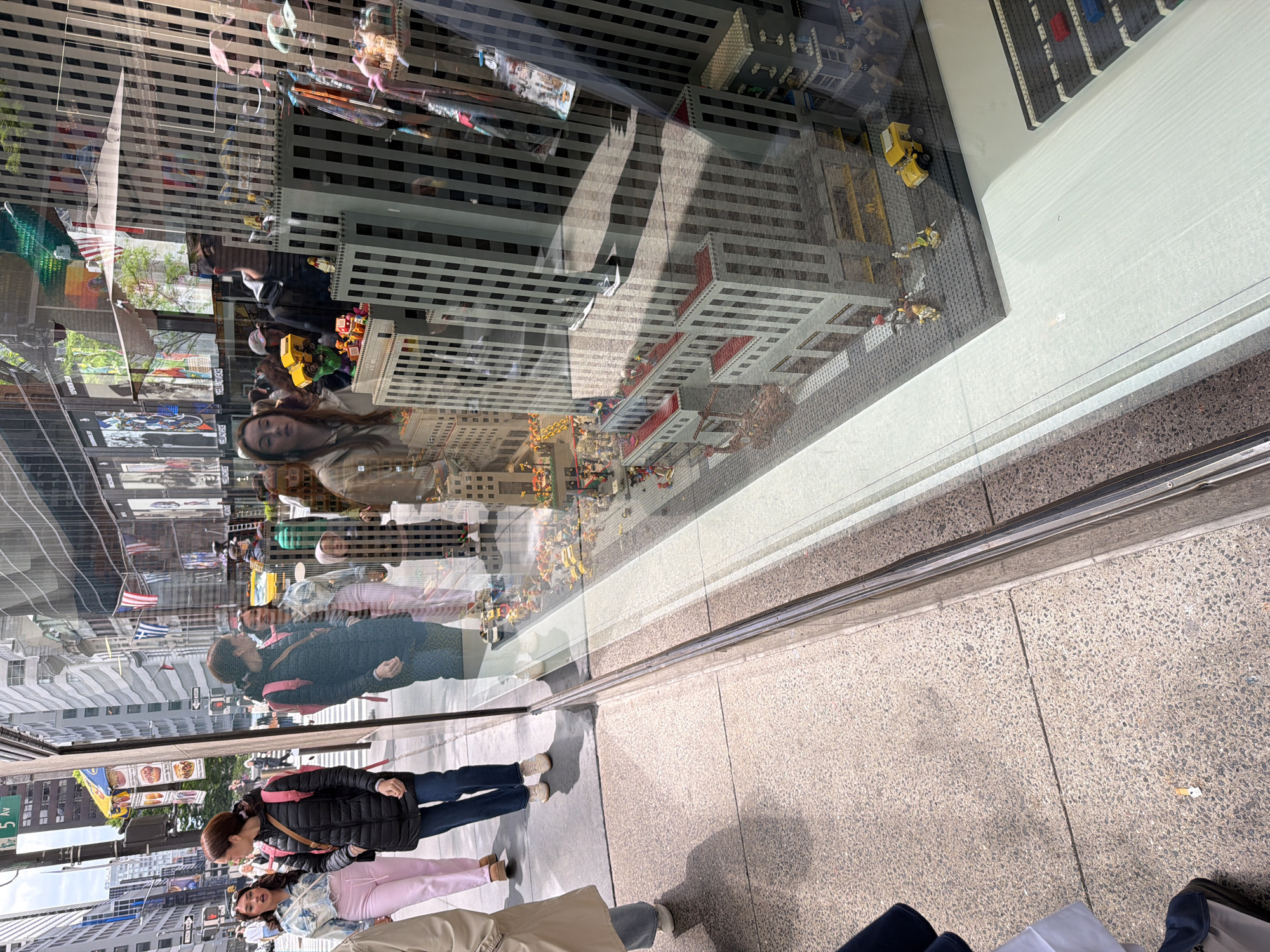 The image shows a scene in New York City, United States of America. There's a storefront window reflecting pedestrians and the street. Among the people reflected are two women; one wearing a pink outfit and another wearing blue jeans. In the window's interior is a detailed Lego model cityscape, featuring skyscrapers and miniature figures.