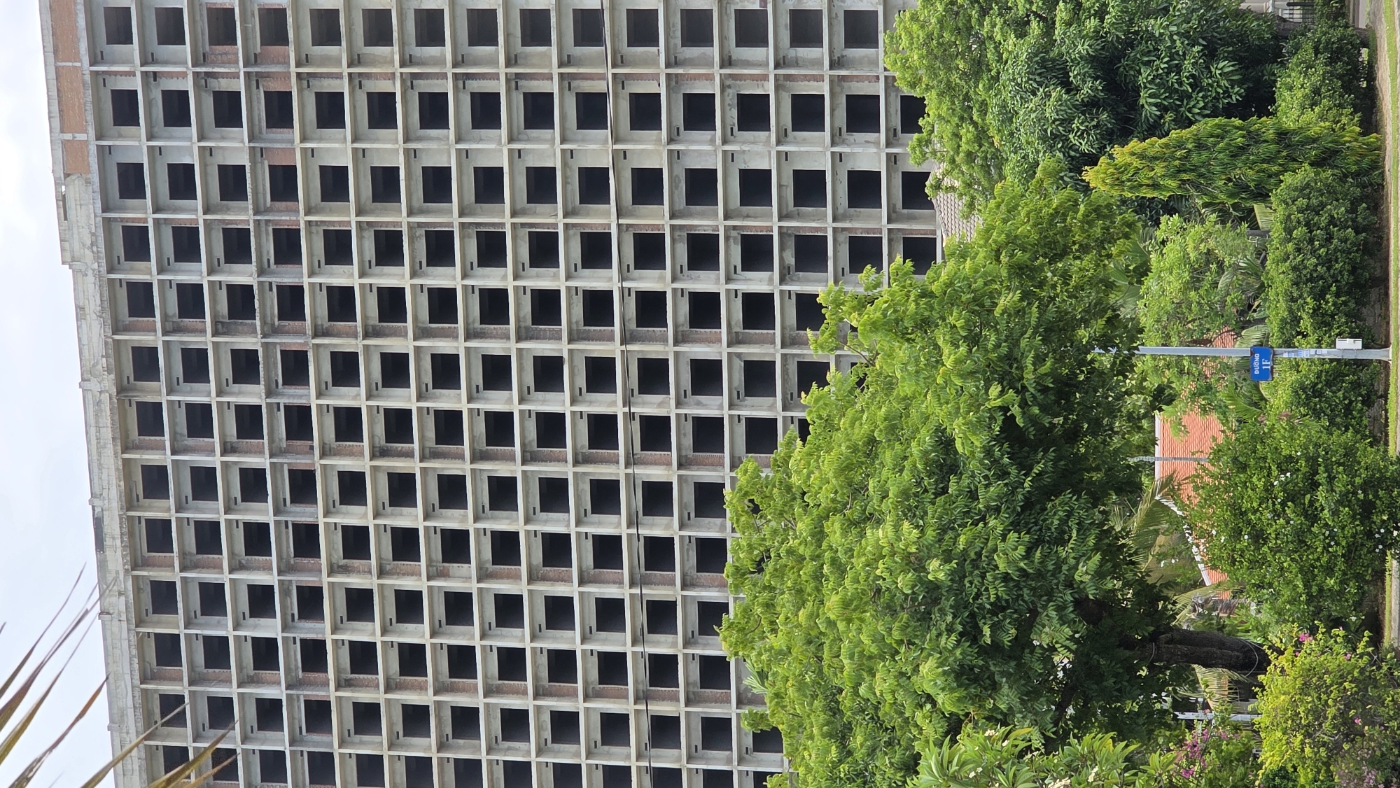 The image shows an unfinished, multi-story concrete building with numerous square window openings. It's adjacent to lush green trees and vegetation. A street sign is visible. The location is Nam Nha Trang Ward, Vietnam. There are no people visible in the image. The focus is on the stark contrast between the architectural structure and the natural environment. The building appears to be abandoned or under construction.