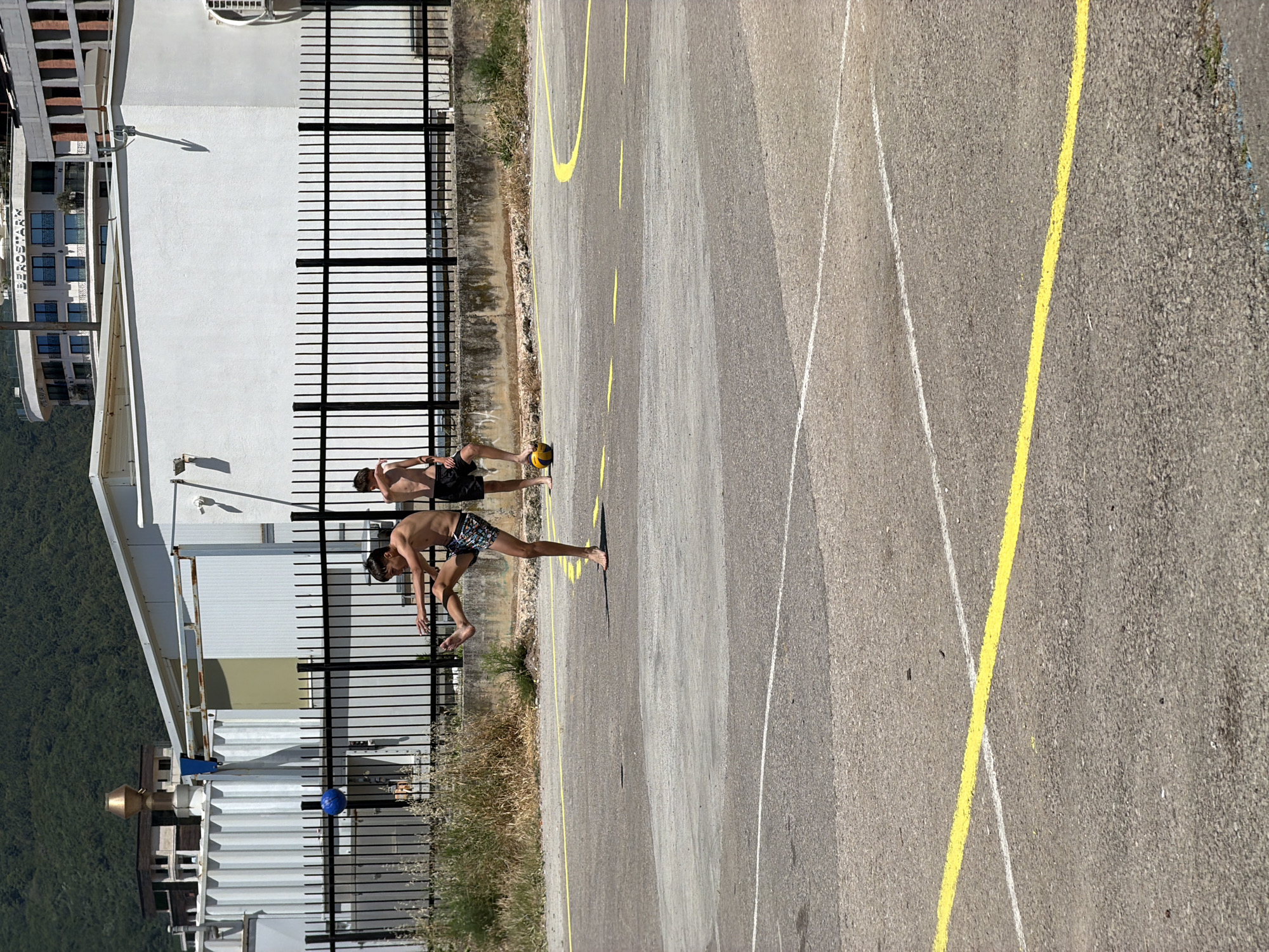 This image depicts two young males playing with balls on a sunlit, asphalt-paved court in Budva, Montenegro. The setting is an outdoor, somewhat neglected area with worn yellow and white lines on the ground.
In the foreground, a young male, bare-chested and wearing patterned swim trunks, is captured mid-action with his left leg extended, appearing to kick an unseen object or performing a maneuver. His right foot is planted firmly on the ground, and he is barefoot. To his right, a second young male, also bare-chested and wearing dark swim shorts, stands with his left foot resting on a yellow volleyball. He is looking towards the first male and has his right hand near his face. Both individuals appear to be in their early to mid-teens.
Behind the playing area, a black metal fence separates the court from a series of white, modular or utilitarian buildings, possibly storage containers or industrial units. Beyond these structures, a low concrete wall with some vegetation is visible. In the upper background, a large, curved, multi-story hotel with balconies, identified by the "BEROSTAR" sign, stands prominently. Adjacent to it, another multi-story building with a more rectangular design is partially visible. A large, green, forested mountain range dominates the far background under a bright sky. A blue ball is noticeable behind the fence, resting against one of the white containers. The scene is brightly lit by direct sunlight, casting shadows.