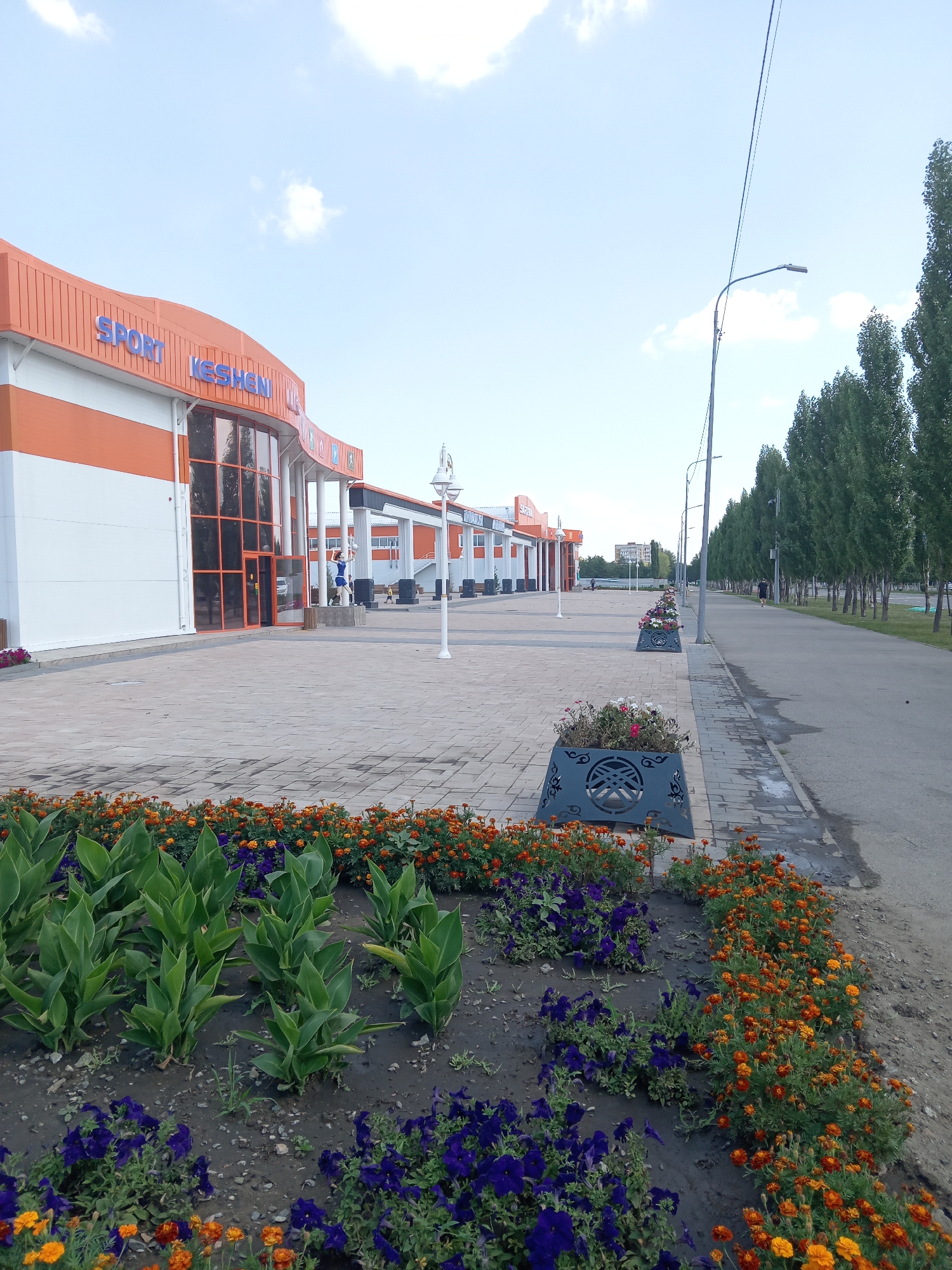 The image shows a modern commercial building with orange and white panels, featuring large glass windows and a prominent sign reading "SPORT KESKEN". In front of the building, there is a wide paved walkway. To the right of the walkway, a street is lined with tall, slender trees. A row of decorative planters containing flowers, predominantly orange and purple, runs along the edge of the walkway. In the foreground, more flowerbeds are visible. In the distance, a street lamp and power lines are present. The sky is partly cloudy. The image is captured in the city of Rudny, Kazakhstan.