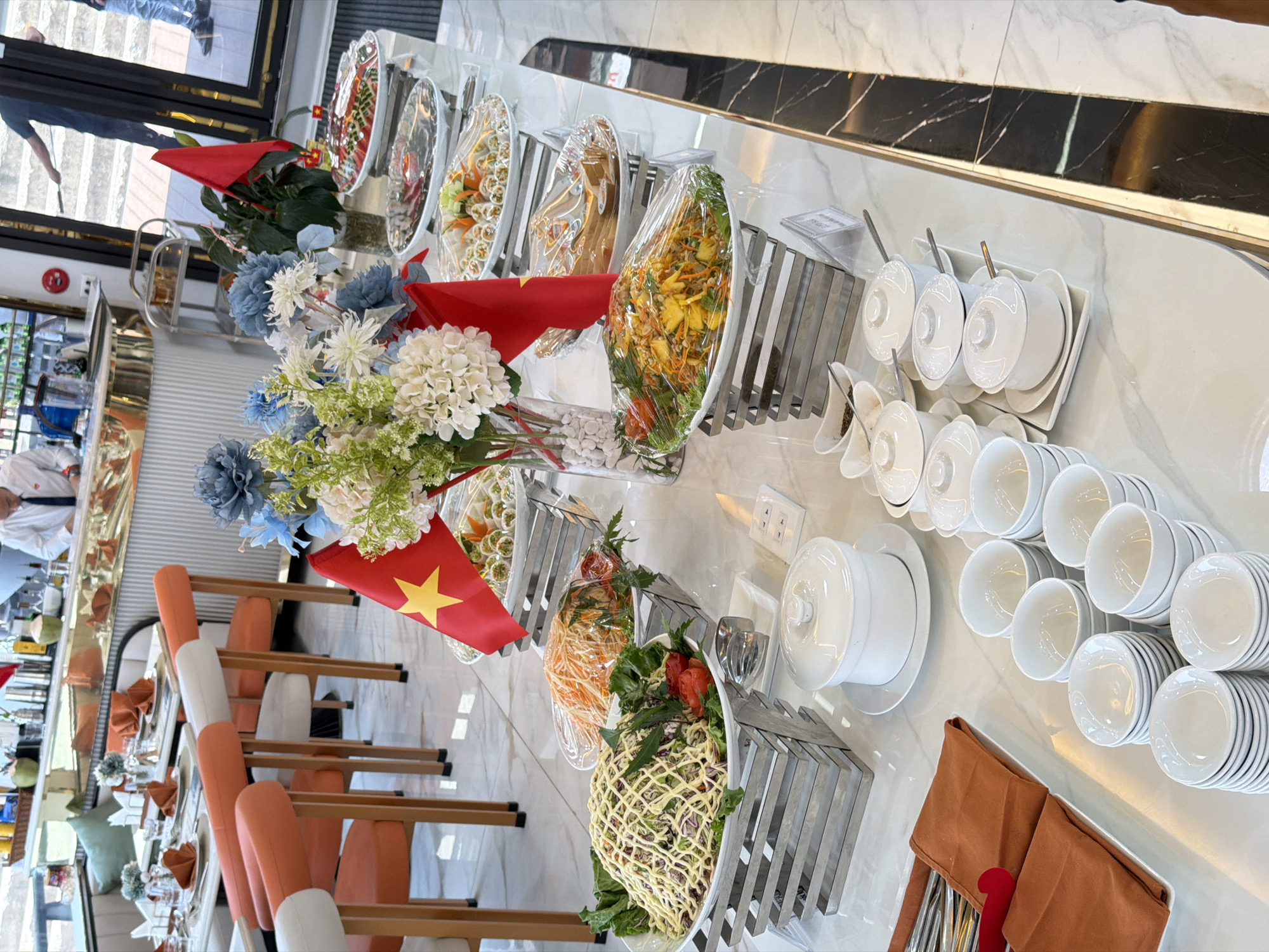 This indoor scene captures a meticulously arranged buffet spread, likely within a hotel or restaurant setting in Tuan Chau Ward, Vietnam. The central feature is a long, gleaming white buffet table with a marble-like surface, showcasing a variety of dishes.
Numerous oval-shaped serving platters hold different salads and prepared food items, many covered with clear plastic wrap to preserve freshness. These dishes display colorful ingredients such as shredded carrots, green leafy vegetables, and other mixed components. Smaller, individual portions, possibly spring rolls, are also visible.
Adjacent to the food, stacks of clean, white ceramic bowls, plates, and lidded serving tureens are neatly organized for guest use. Cutlery, including spoons and forks, is presented in tidy burnt orange fabric pouches.
Decorative elements include elegant artificial floral arrangements featuring blue, white, and green flowers in clear vases. Small Vietnamese flags, identifiable by their red color and yellow star, are strategically placed throughout the buffet, subtly indicating the location.
In the background, to the left, a dining area with orange and white upholstered chairs and tables is partially visible, hinting at the seating arrangements. A staff member in a white shirt can be seen attending to the area, and a reflection of a person's lower body is visible in a polished surface or window, suggesting an active environment.
The overall presentation suggests a formal or semi-formal dining experience, with the buffet prepared for a meal service. The scene is orderly and well-lit, though the specific time of day is not discernible. No prominent text is visible on any signs or labels.