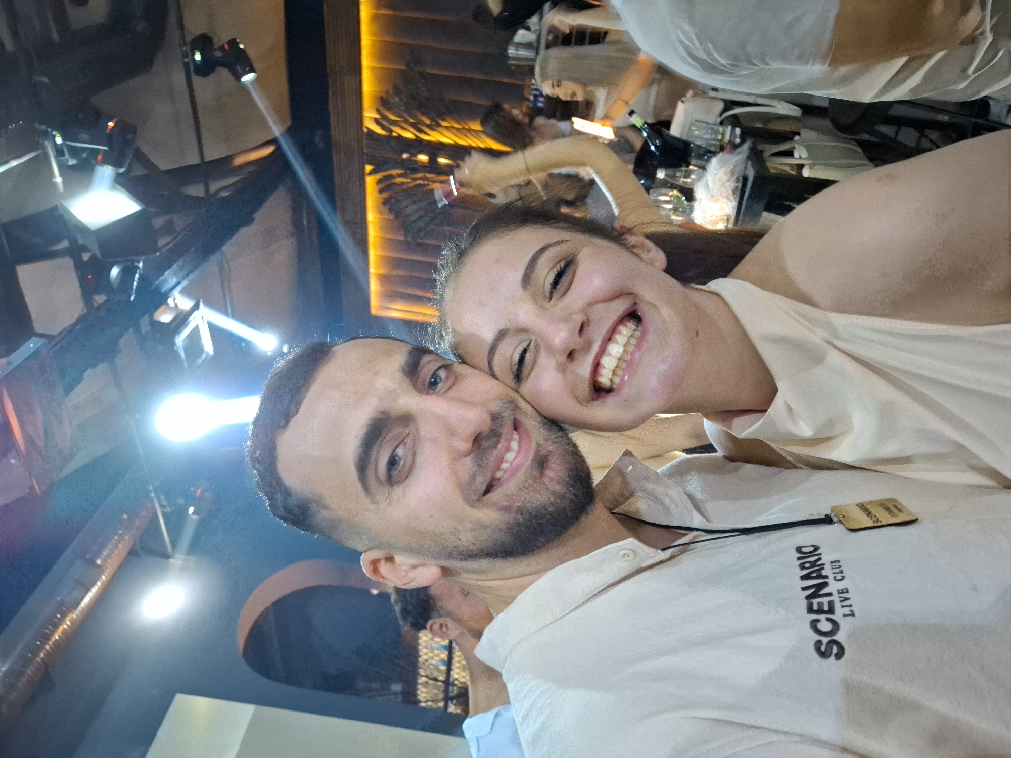 The image depicts a close-up, selfie-style photograph taken inside a nightclub or live music venue identified as "Scenario Live Club" in Tsarevo, Bulgaria.
In the foreground, a male and a female subject are positioned closely, smiling at the camera. The male, positioned to the left, has dark hair, a beard, and blue eyes. He wears a light beige or off-white collared shirt with "SCENARIO LIVE CLUB" embroidered in dark letters on the left chest area. A dark lanyard with a wooden tag, partially inscribed "SCENARIO," hangs around his neck. The female, positioned to the right and leaning into the male, has dark brown hair and is smiling broadly, revealing her teeth. She wears a light beige or off-white halter-neck top. Both individuals appear to be in their twenties or early thirties.
The background reveals a dimly lit club environment with artificial lighting. Above the subjects, the ceiling features exposed ductwork and several bright spotlights, including moving head fixtures, projecting beams of light across the space. To the far left, a dark wall with an arched opening is visible. To the right, behind the female, a decorative wall is illuminated with warm orange backlighting, featuring vertical panels and what appears to be a stylized dark, tree-like sculptural element.
Several other individuals are visible in the mid-ground and background. One female figure is seen raising her right arm, holding a small red object or cup. Another female with blonde hair is visible further right, appearing to hold a mobile phone. A table or bar area is present in the mid-ground, displaying bottles (including one dark bottle, possibly an alcoholic beverage), glasses, and other items. The overall scene suggests a lively social gathering within the club setting.