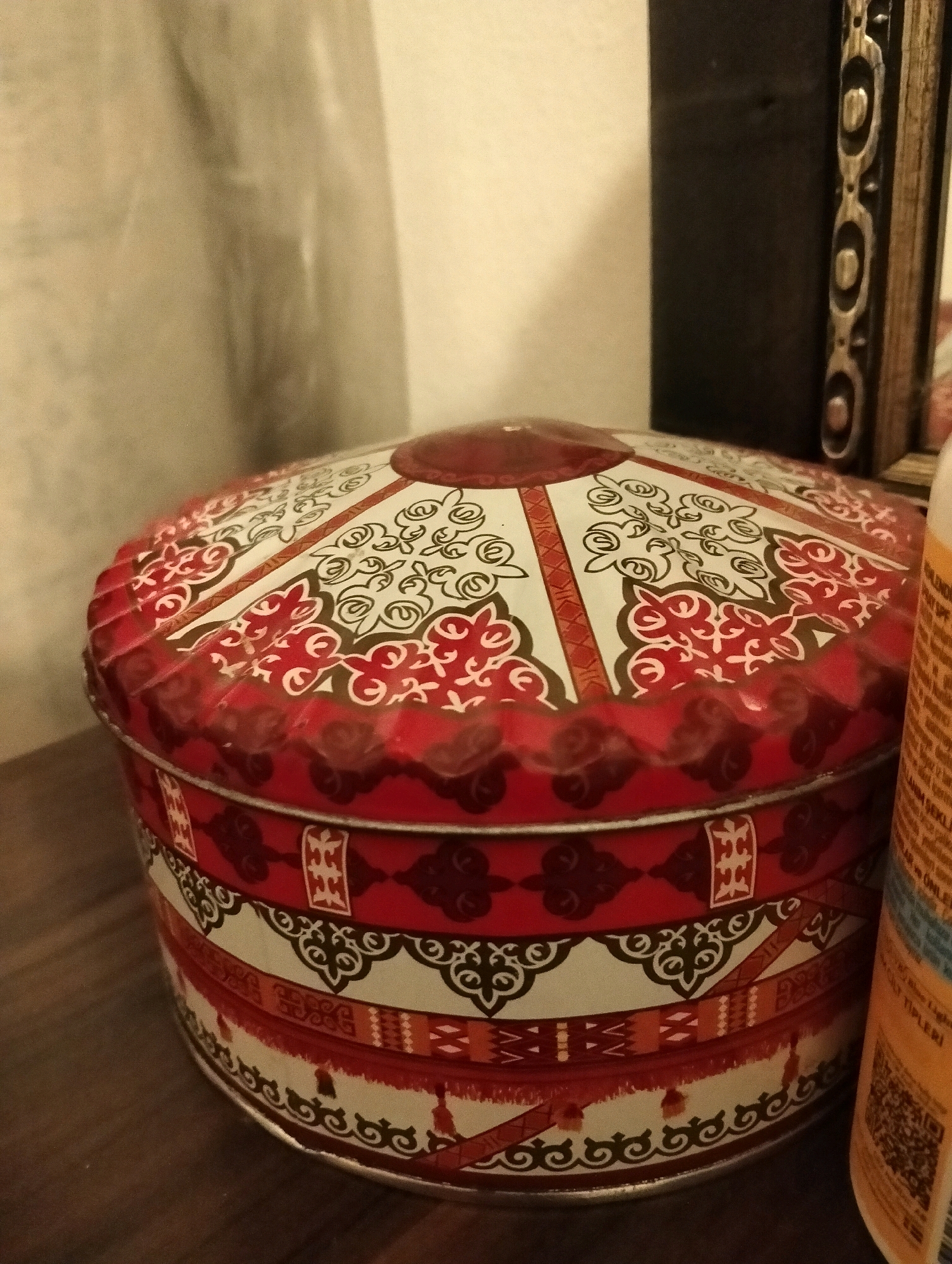The image shows a close-up of a decorative tin container with a lid, featuring a red and white patterned design that appears to be inspired by traditional motifs. The container is round and has a slightly raised, scalloped edge around the lid. The pattern on the lid includes intricate swirling lines and geometric shapes in red and black against a white background, bordered by red sections. The side of the container also displays bands of red, white, and darker red with various patterns.
The tin is placed on a wooden surface, possibly a table or shelf. In the background, to the right of the tin, a portion of a yellow label with text and a QR code is visible, likely belonging to another object not fully in frame. To the left and behind the tin, a pale, textured surface is present, possibly a wall or drapery, with some shadows creating a diffused lighting effect. To the far right and slightly behind the tin, a dark, rectangular object with a decorative edge is visible, which could be a picture frame or a piece of furniture. The lighting is subdued, casting soft shadows and highlighting the textures of the objects. The overall impression is a still-life arrangement on a domestic surface, with no people or explicit urban elements visible to indicate a specific location in Balçova, Turkey.