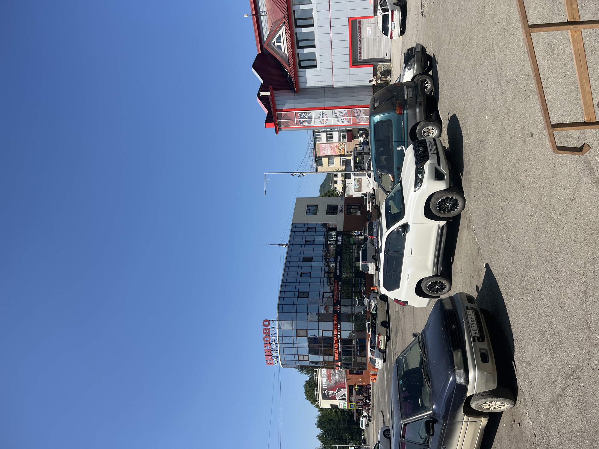 The image depicts a street scene in Yelizovo, Russia, under a clear blue sky. Several vehicles are parked in a lot or on the street, including a dark blue minivan in the foreground, a white SUV, and a green SUV. In the background, there is a multi-story building with a curved glass facade, featuring red lettering that appears to read "БЛИЗОВО". Adjacent to this is another building with a red roof and grey siding, displaying signs and an open garage door. A person is visible standing near the grey-sided building. The environment suggests a commercial or business district, with other buildings and trees visible further in the background. The overall impression is one of a typical sunny day in a Russian town.