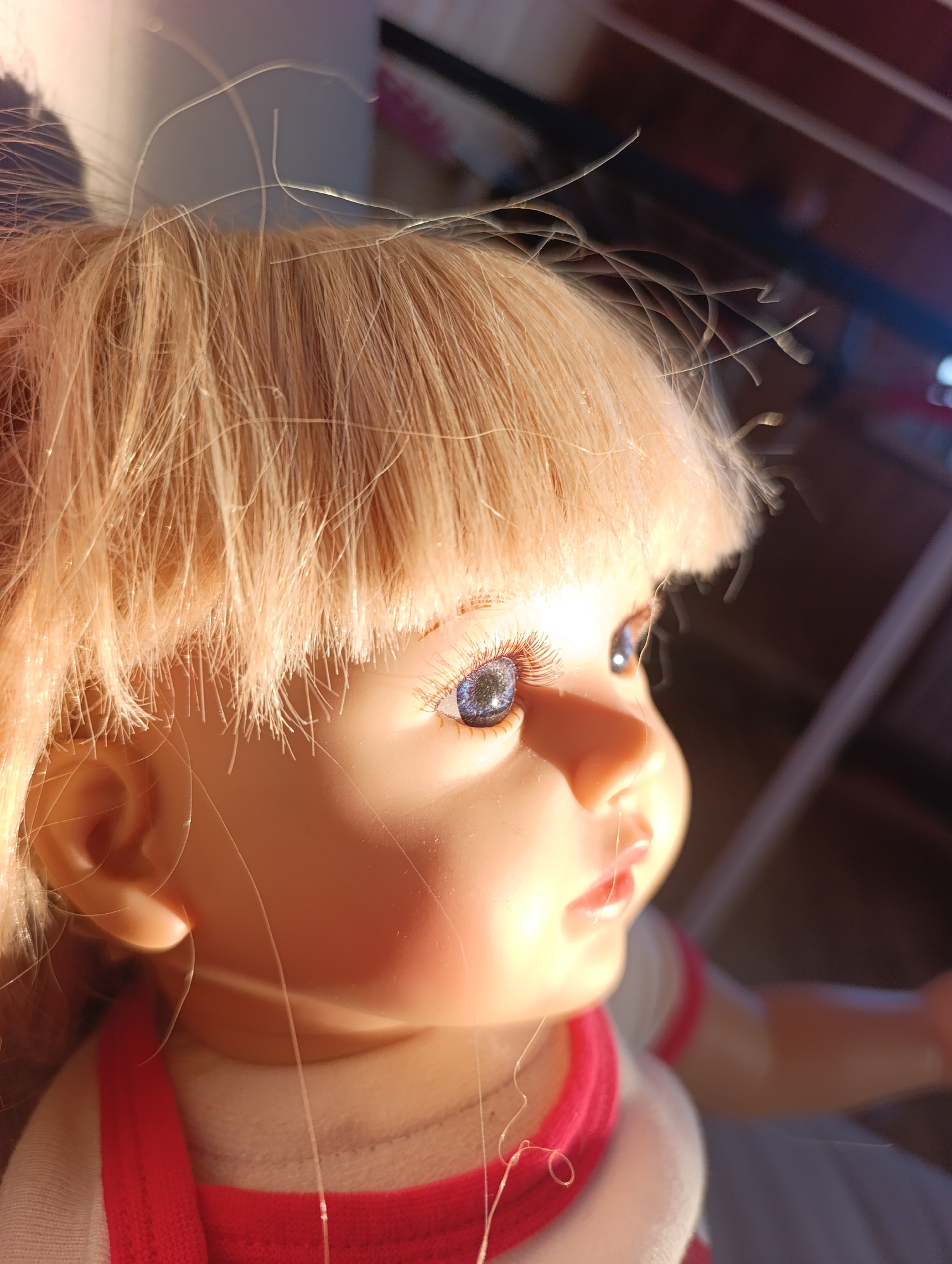 A close-up shot depicts the profile of a doll with blonde hair, styled in a short, blunt cut. The doll has blue eyes with long eyelashes. Its skin tone is light, and its facial features are well-defined, including a small nose and slightly parted lips. The doll is wearing a white shirt with red trim around the neckline. Thin, translucent fibers are visible on the doll's face and hair. The background is blurred, but suggests an indoor setting. There is no indication of the location being Krasnoyarsk, Russia.