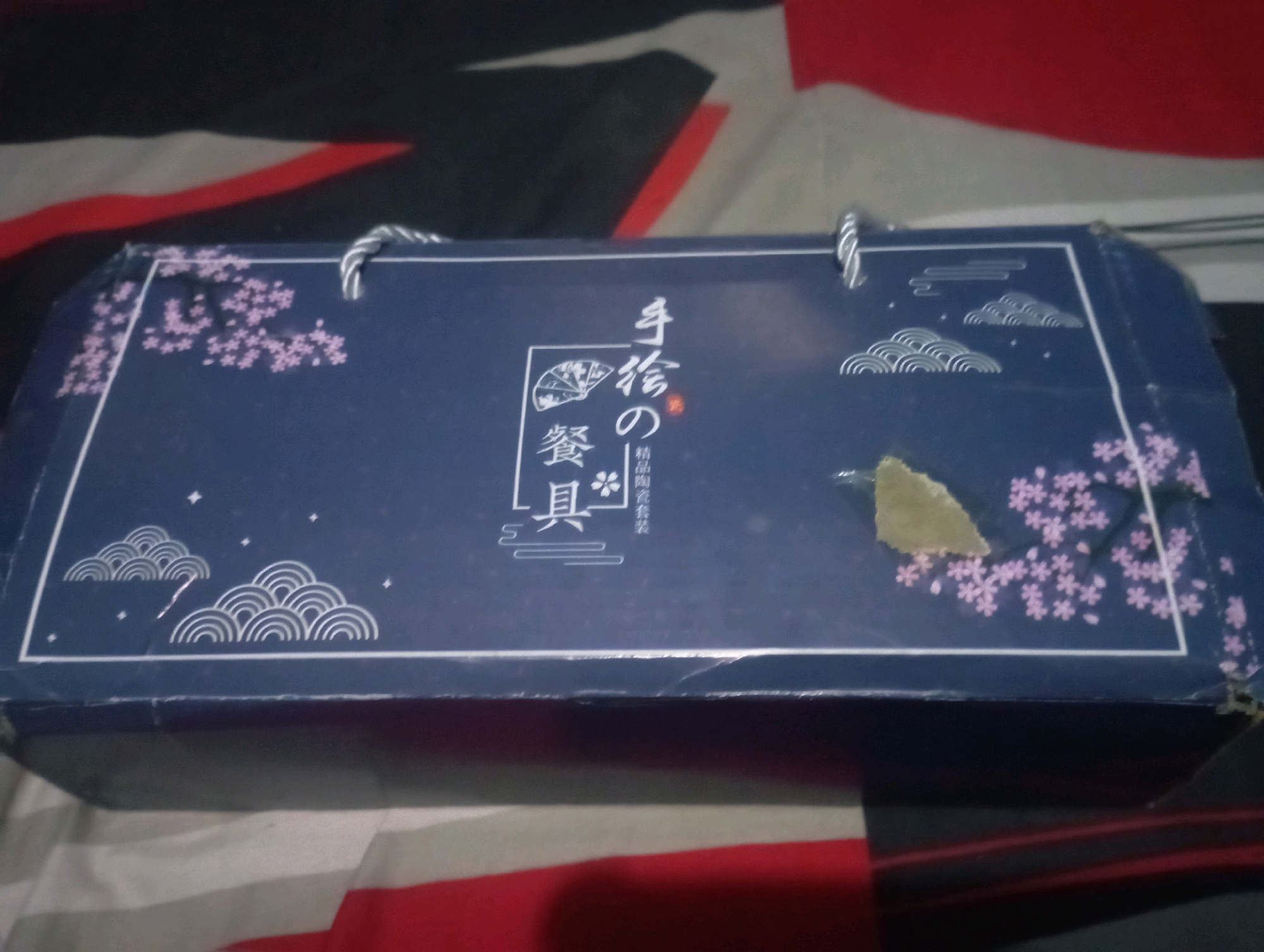 An oblong, dark blue cardboard box with a rope handle on each side rests on a surface with a black, red, and white striped pattern. The box is adorned with various decorative elements. On the left side, there is an illustration of cherry blossoms in light pink and stylized clouds in white. In the center, there is a rectangular panel with Japanese characters. To the right of this panel, there are more illustrations of stylized clouds in white and scattered cherry blossoms. The box appears to be a product packaging. There is a torn piece of light beige paper stuck to the top right portion of the box. The overall scene is likely indoors, possibly in a domestic setting in Senangsari, Indonesia, though no specific city landmarks are visible.