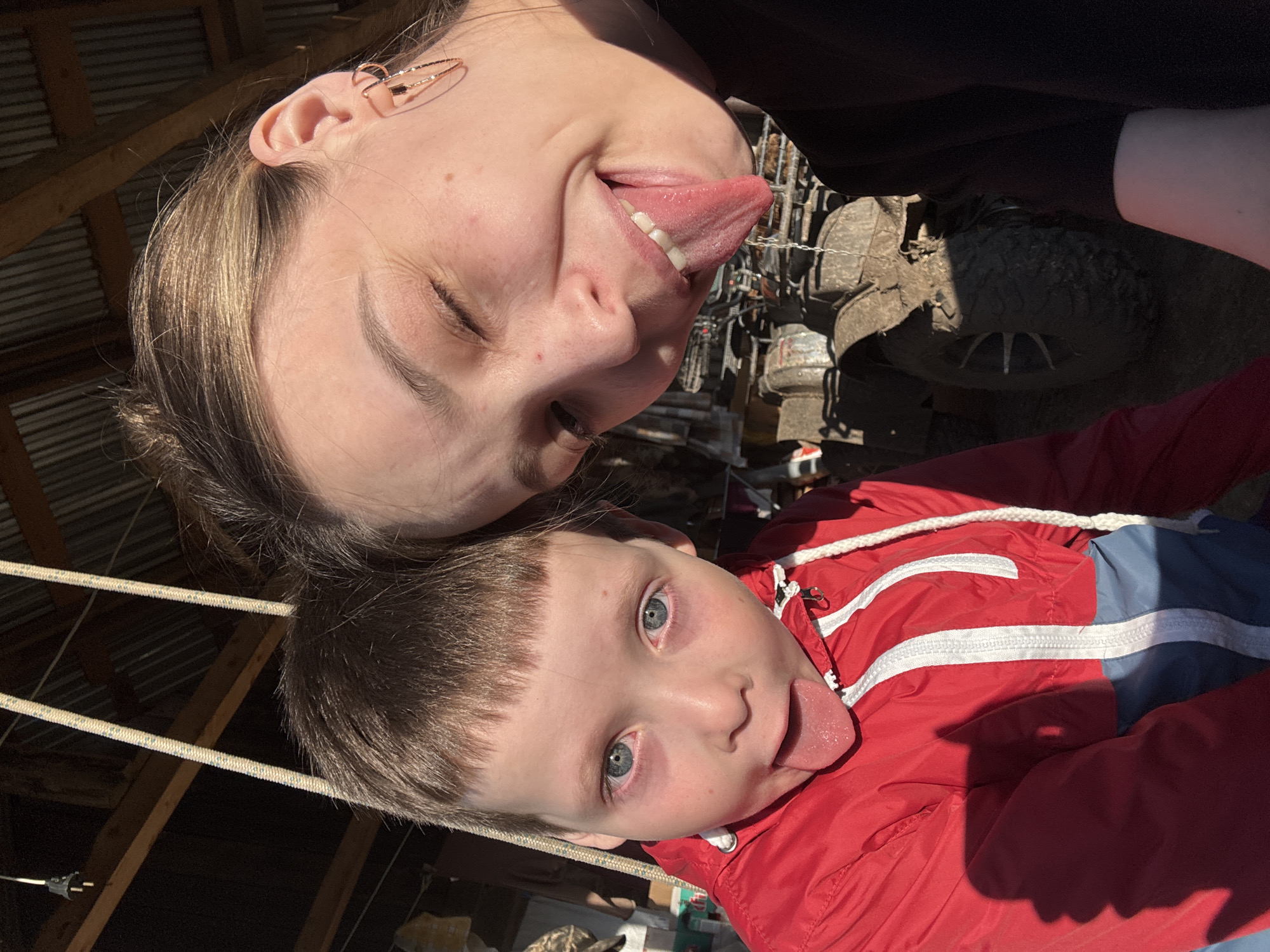 Two individuals, a woman and a child, are posing for a photo indoors, possibly in a garage or barn-like structure. The woman, with blonde hair and an ear cuff, is on top and sticking her tongue out with her eyes closed. The child, with short brown hair and blue eyes, is below her, also sticking his tongue out. The child wears a red and blue jacket. A section of a muddy tire and a piece of machinery are visible in the background. Ropes and wooden beams suggest the structure's nature. There is no definitive evidence to confirm a location in Кемуль, Russia.