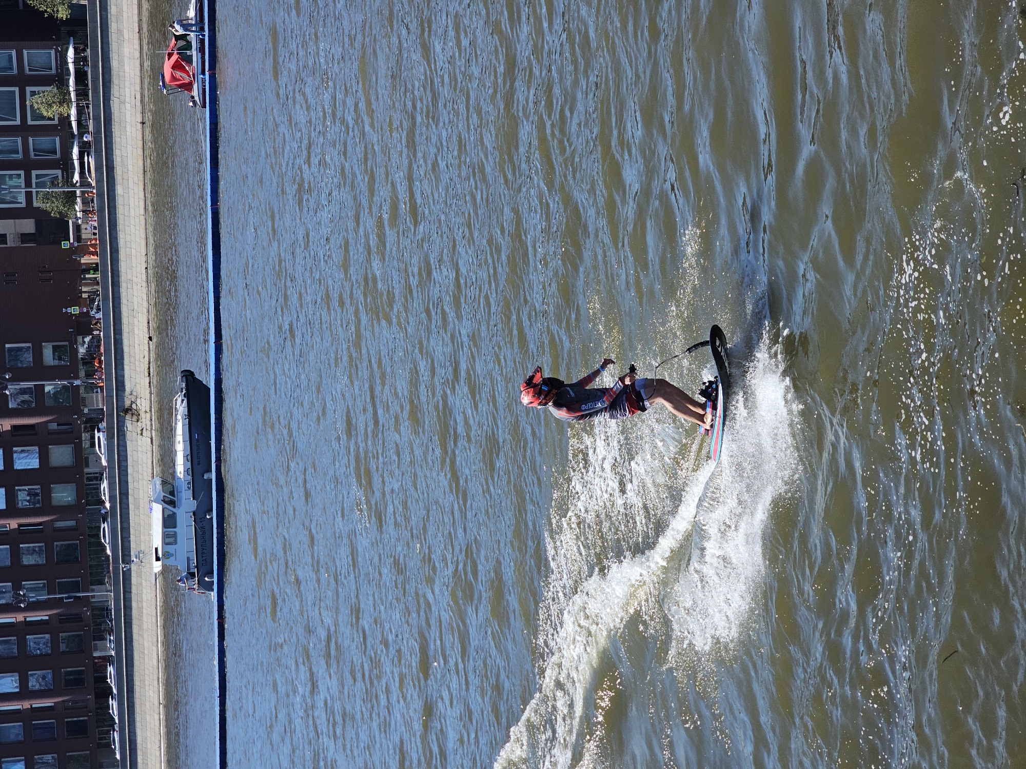 A person is riding a jet board on a body of water, creating a wake. They are wearing a red helmet and a red and dark-colored athletic outfit. The person is positioned on the right side of the frame. In the background, there is a large, multi-story building with many windows, characteristic of urban architecture. In the middle ground, there is a body of water with a blue floating barrier. Two boats are visible on the water, one is a patrol boat with the inscription "ПАТРУЛЬНЫЙ-15" (Patrol-15) and the other has a red canopy and a person standing on it. The scene appears to be a sunny day in Moscow, Russia, possibly on the Moskva River.