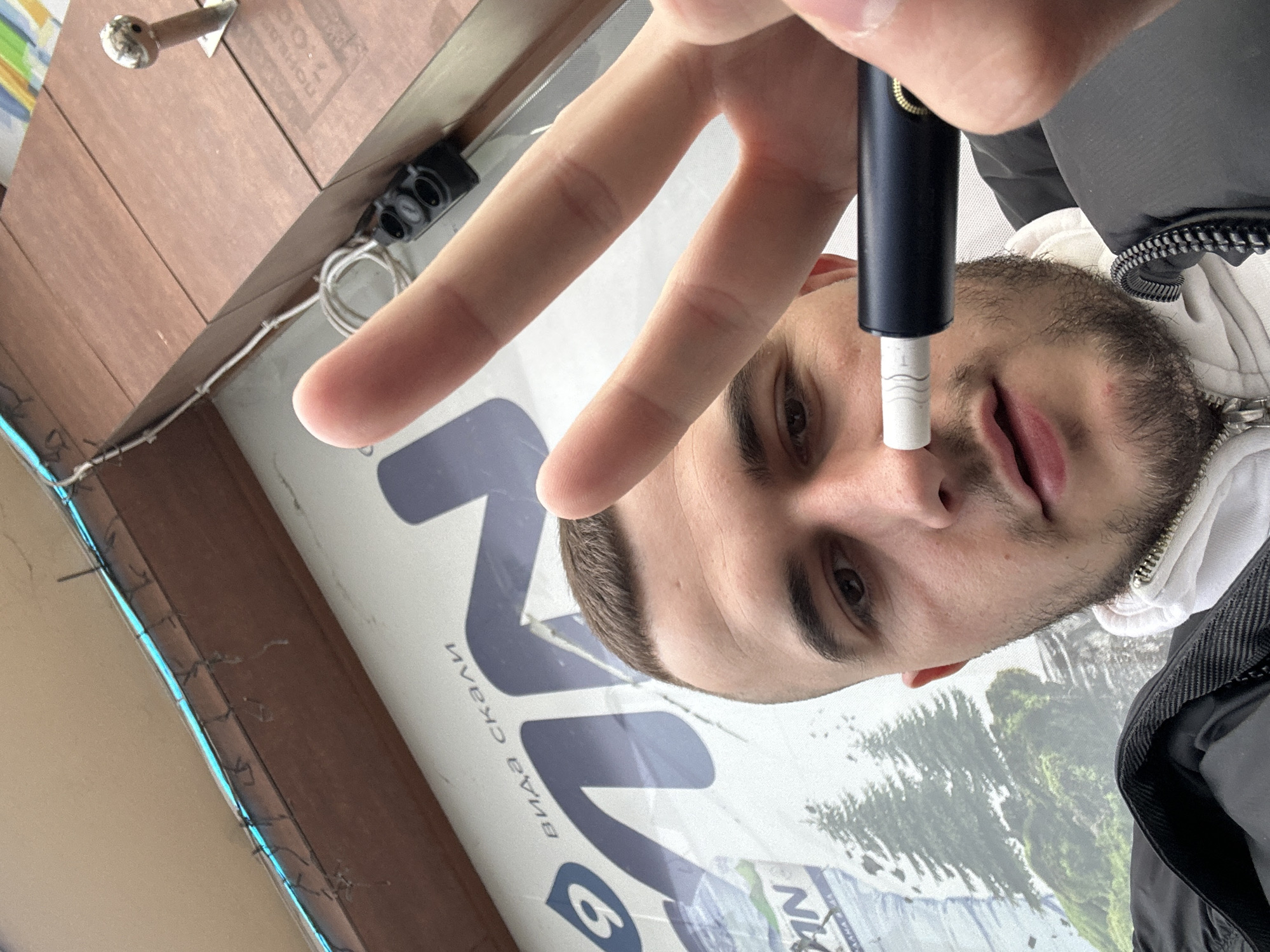 A man poses with a vaporizing device under his nose, framed by a peace sign with his fingers. He is standing in front of a large advertisement featuring the logo for "Uvex," a ski equipment brand, and the partially obscured text "виа скали." This suggests the advertisement is likely for a store selling winter sports equipment in Plovdiv, Bulgaria, although further visual confirmation of the location within Plovdiv cannot be determined from the available information. The man wears a black jacket over a white hooded sweatshirt. Part of the surrounding architecture is visible, including tiling, exposed wiring, and a nearby electrical outlet.
