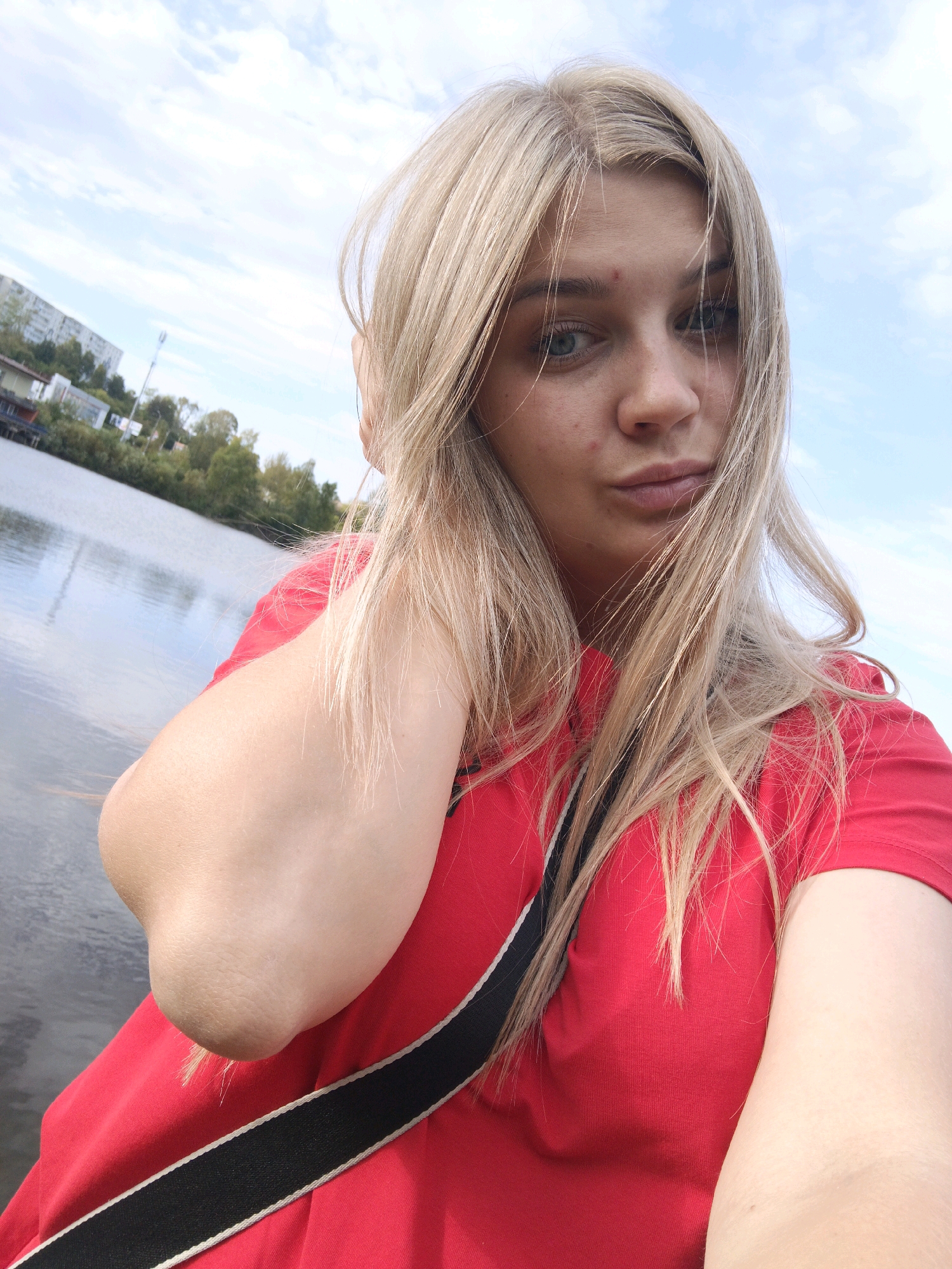 A young woman with blonde hair and a red shirt stands by a body of water under a cloudy sky. She is looking at the camera and has her arm raised, holding her hair. In the background, there are trees, buildings, and a cellular tower, suggesting an urban or suburban setting. The water appears to be a lake or a wide river. The overall scene is casual and appears to be a selfie taken outdoors. Without further information, it is not possible to pinpoint the exact location within Zlatoust, Russia.