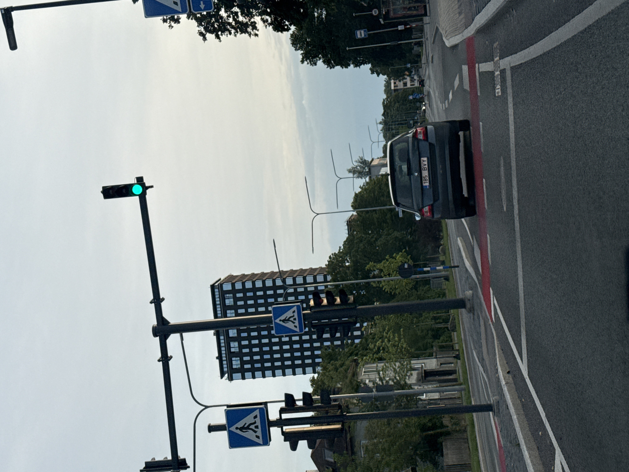 The image shows a street scene in Tallinn, Estonia, with a dark-colored car driving away from the camera on a multi-lane road. The car has the license plate "185 BVY". The road has clear lane markings, including a red-painted pedestrian crossing. Traffic lights are visible, with one showing a green light. Pedestrian crossing signs are also present. In the background, a tall, modern building with a dark facade and many windows stands out, along with trees and other buildings. The sky is overcast.