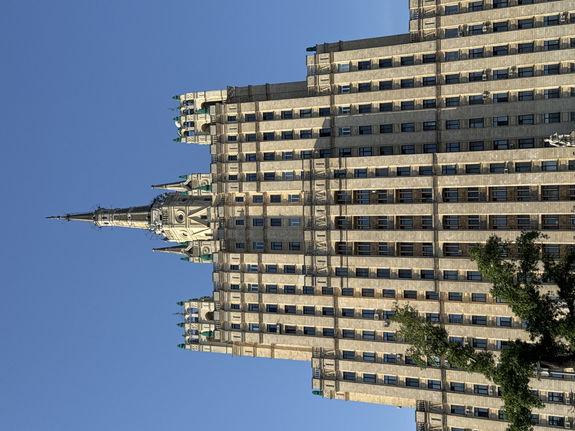 The image shows the Ministry of Foreign Affairs building, one of the "Seven Sisters" skyscrapers located in Moscow, Russia. The building is captured from a low-angle perspective. The architectural features include the multi-story structure, numerous windows, and the spire at the top. The sky is clear and blue. A tree is partially visible in the foreground on the right.