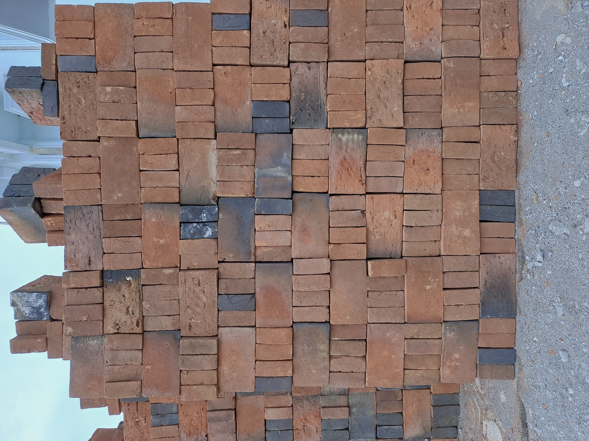 A large stack of red and grey bricks is piled high against a light-colored building facade in Lhokawe Teungoh, Indonesia. The bricks are of varying sizes and conditions, with some appearing worn and chipped. They are stacked in an uneven, somewhat haphazard manner, with several rows reaching well above eye level. The ground beneath the stack is covered in gravel and small stones. The sky is visible at the top of the frame, appearing pale blue and overcast. The scene depicts a construction or storage area where building materials are being kept.