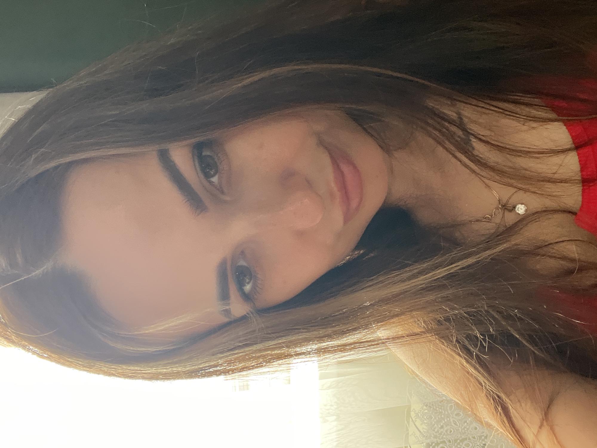 A medium close-up selfie of a woman is captured against a soft, natural light source emanating from the left. The woman has long, brown hair with lighter streaks, which frames her face. Her eyes are dark, and her eyebrows are well-defined. She has a light dusting of freckles across her nose and cheeks. Her lips are slightly parted, and she appears to be smiling subtly. She is wearing a red top and a delicate necklace with a pendant. The background on the left is a blurred sheer curtain with light filtering through, suggesting an indoor setting near a window. The right side of the background is a darker, solid color, possibly a wall.