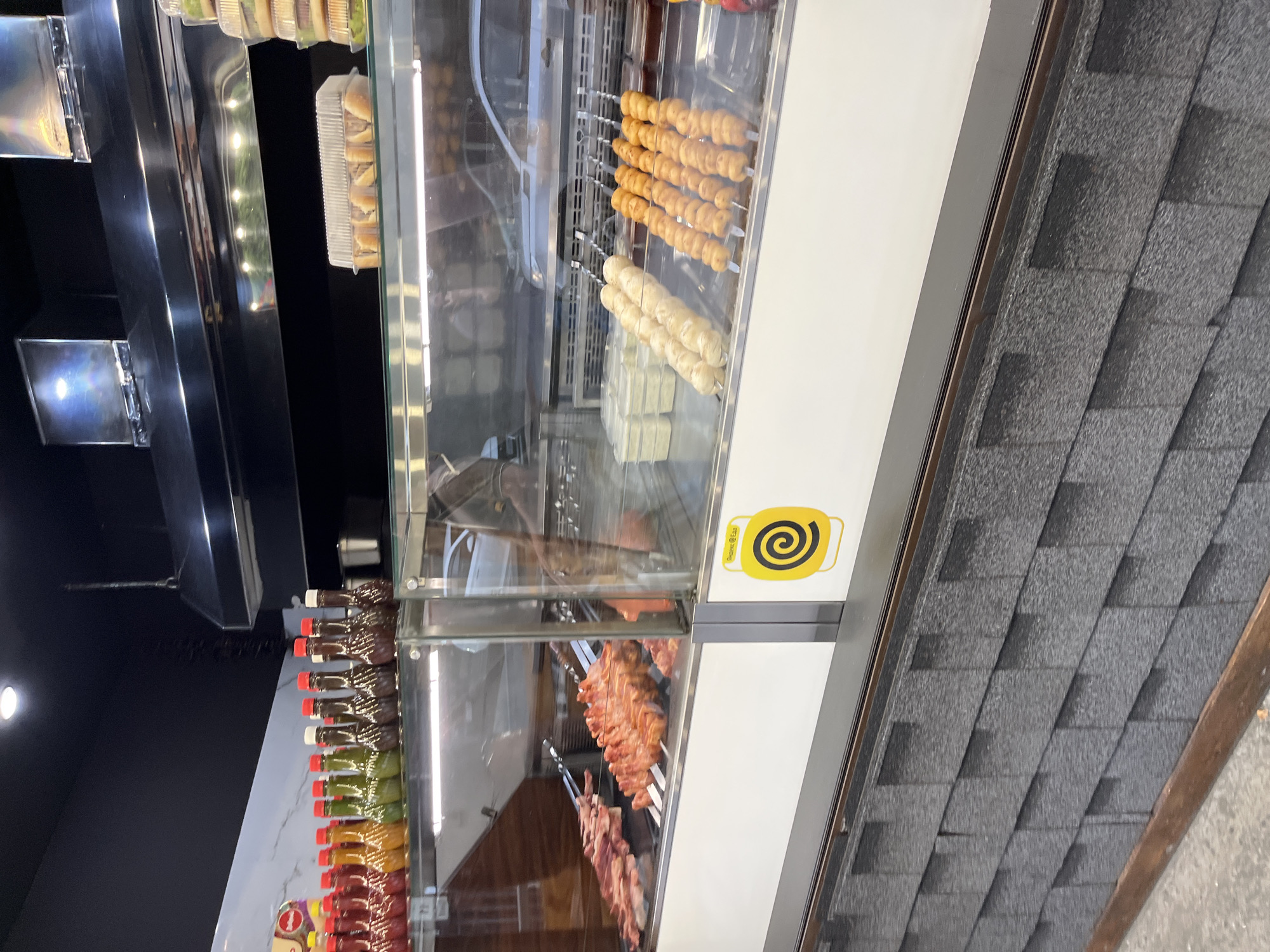 The image shows the interior of a food establishment, likely a cafe or restaurant, with a refrigerated display case. The display case contains various food items. On the left side, raw meat on skewers is visible, suggesting a barbecue or grill-focused menu. To the right, there are rows of skewers with what appear to be cooked meatballs or dumplings. Behind these, clear containers hold a white, possibly creamy, substance. Above the display case, shelves hold bottles of sauces or condiments. The establishment is decorated with a dark ceiling and a patterned tiled wall. A prominent yellow sticker with a black spiral design is affixed to the lower right side of the display case. The overall lighting suggests an indoor setting with artificial lights. The scene appears to be in Sochi, Russia, based on context provided.